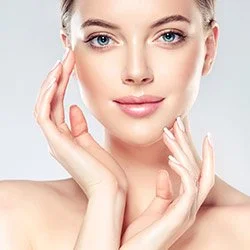 Close-up of a young woman with clear skin, smiling gently, touching her face with both hands.
