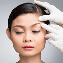 A woman with closed eyes getting a cosmetic or medical procedure on her forehead from a professional wearing white gloves.