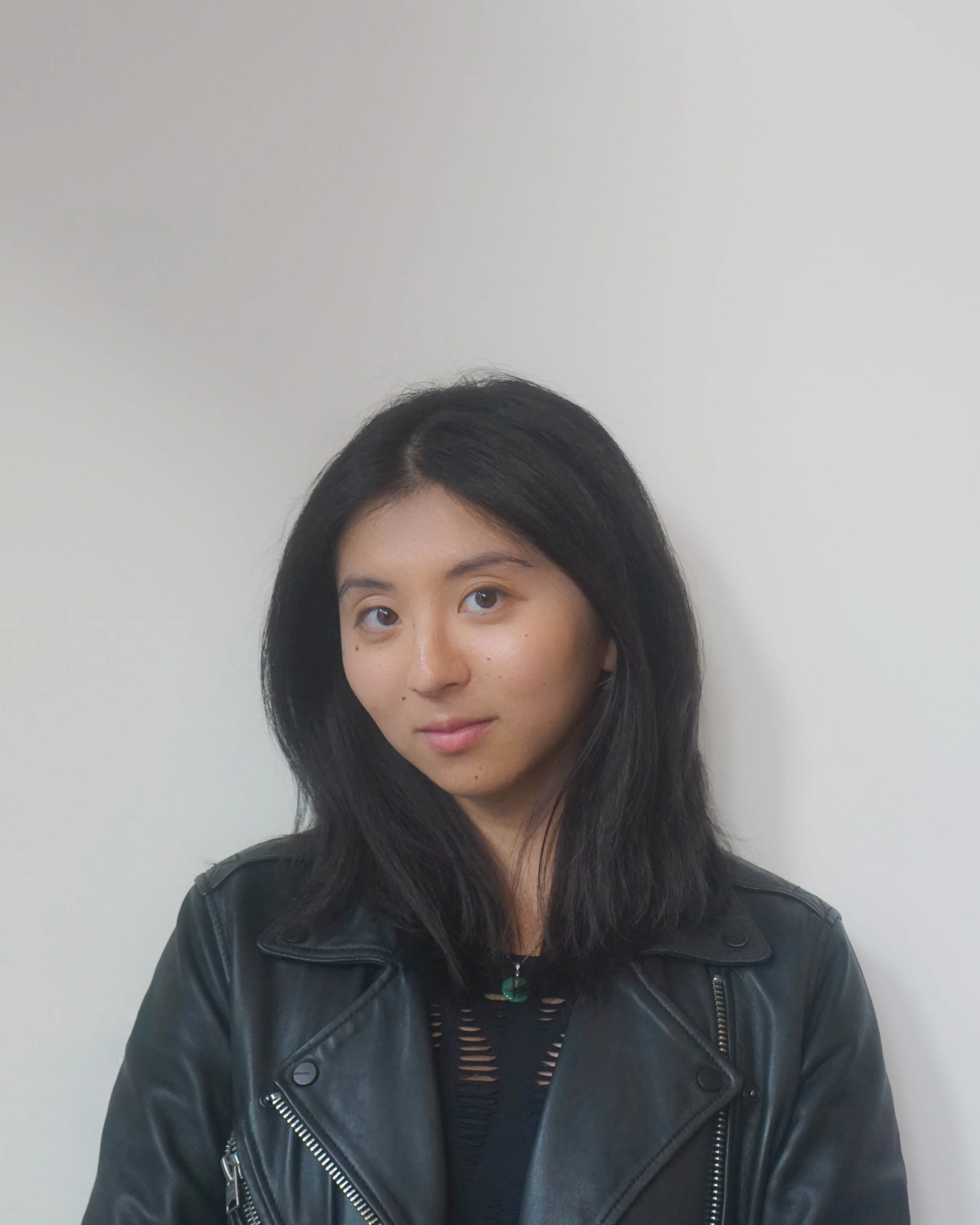 A woman with long black hair, wearing a black leather jacket and a black top, standing against a plain white wall.