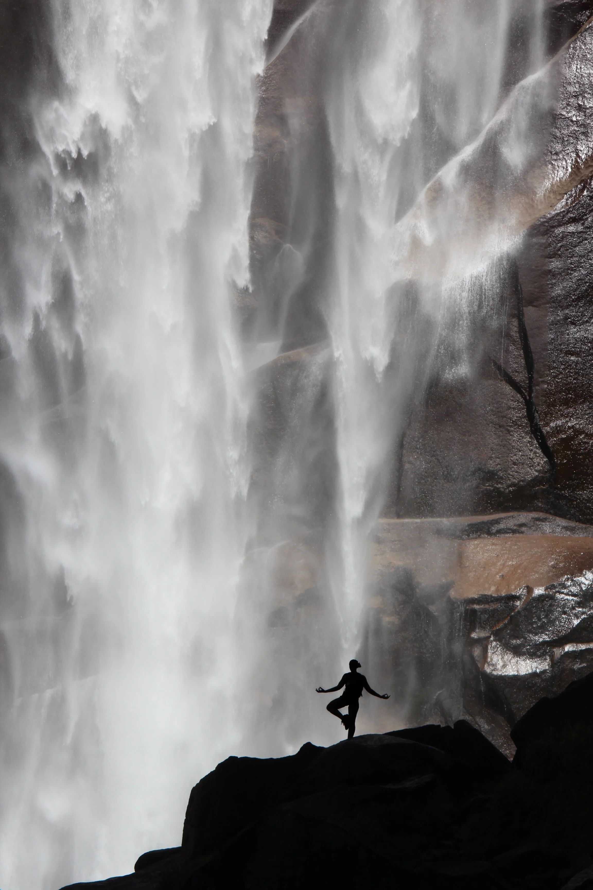 A person standing on a rock in front of a large waterfall, silhouetted against the mist and flowing water.