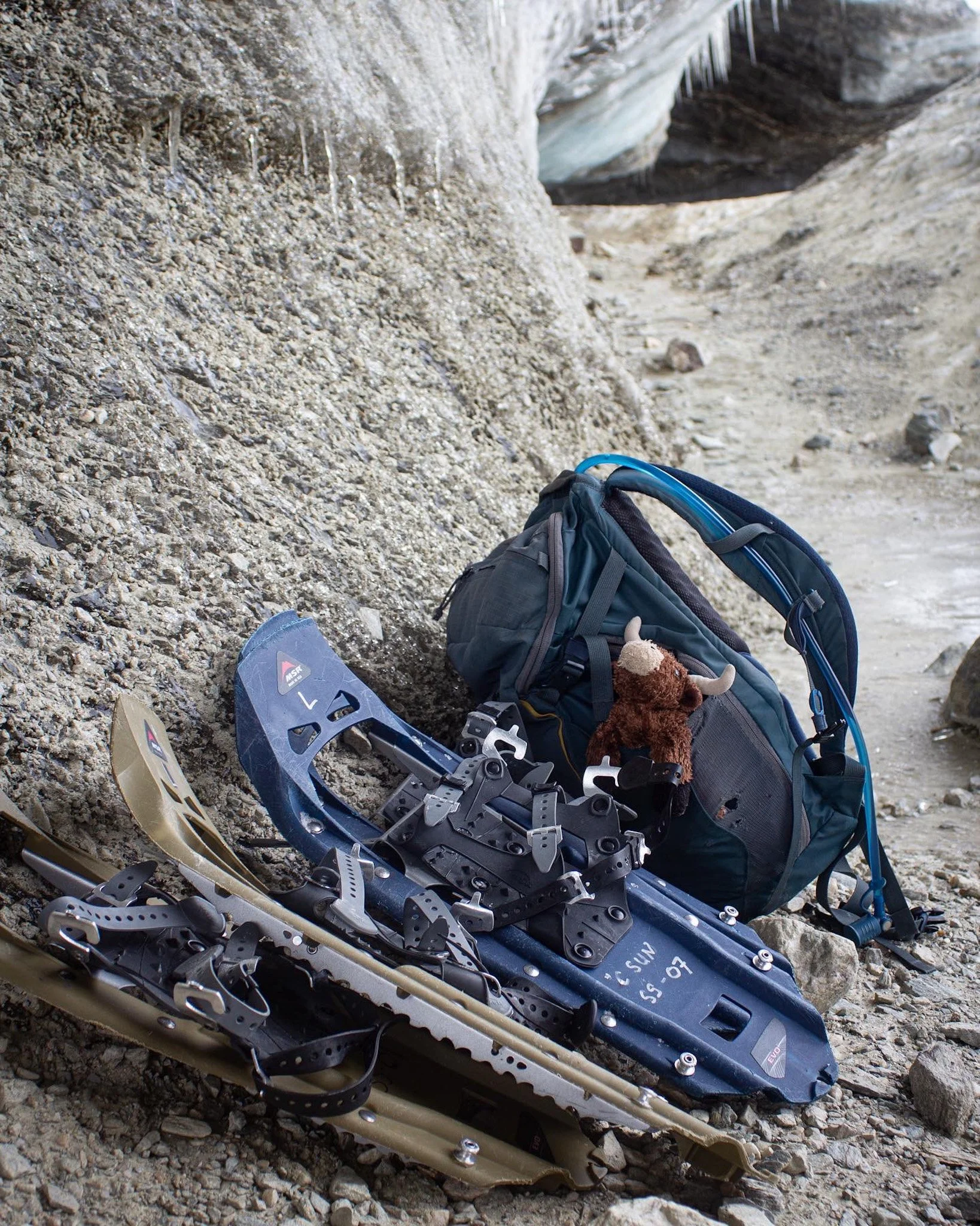 Climbing gear, including skis, a backpack, and a stuffed animal, resting against a rocky mountain trail.