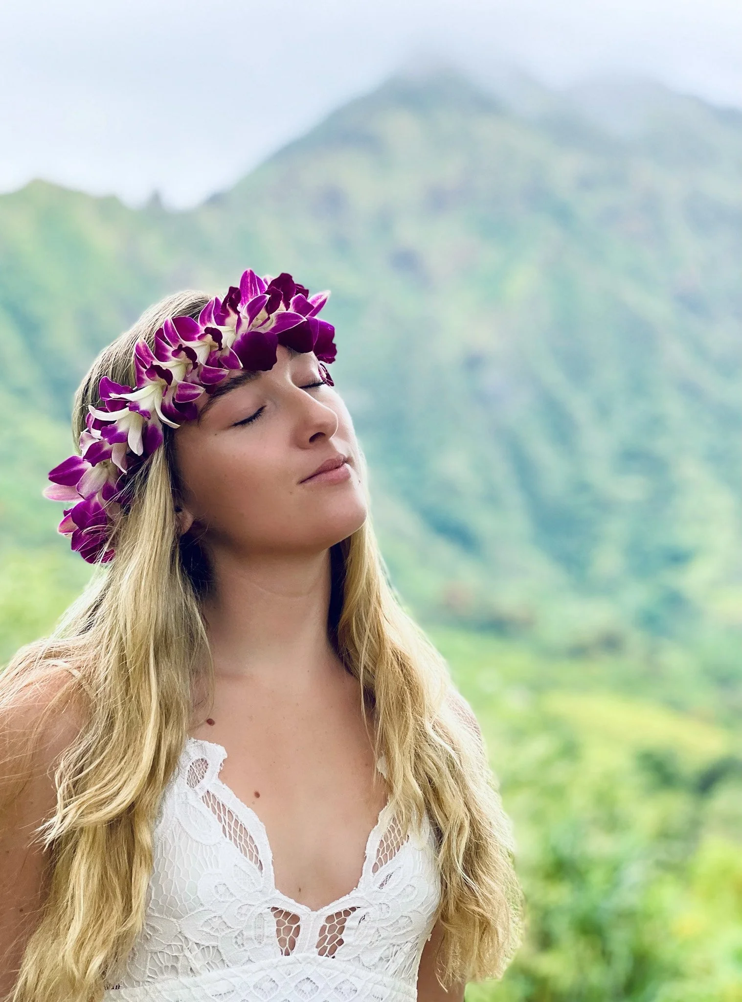 A woman with long blonde hair wearing a white lace dress and a purple and white flower crown, standing outdoors with lush green mountains and cloudy sky in the background, eyes closed.