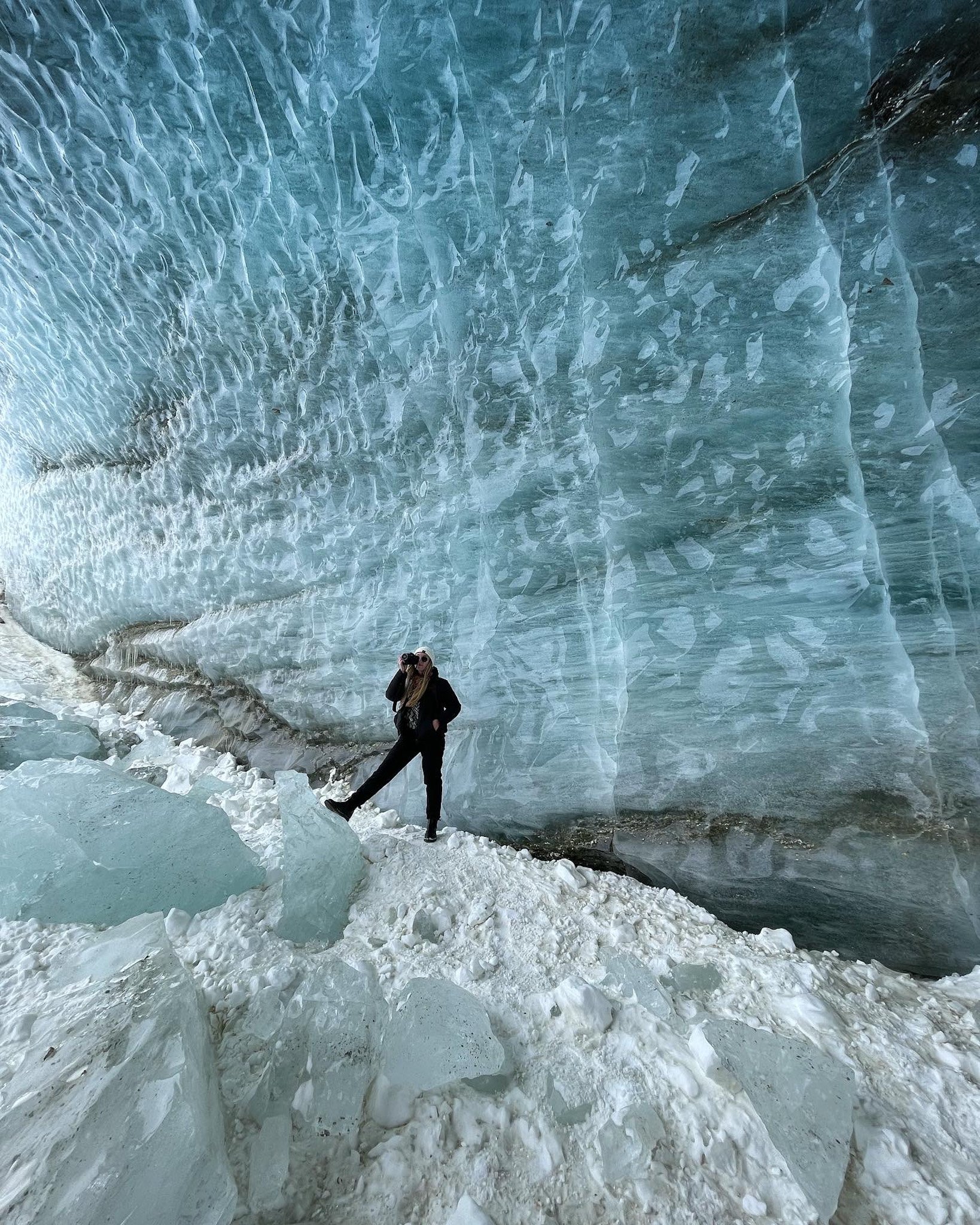 A woman standing on snowy ice next to a large icy blue glacier formation.