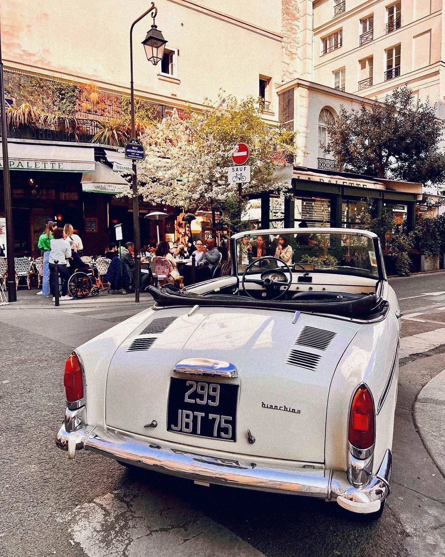 A vintage white convertible car parked on a city street in front of a café with outdoor seating, with people dining, trees, and apartment buildings in the background. There are street signs including a 'no entry' sign and a street name sign.