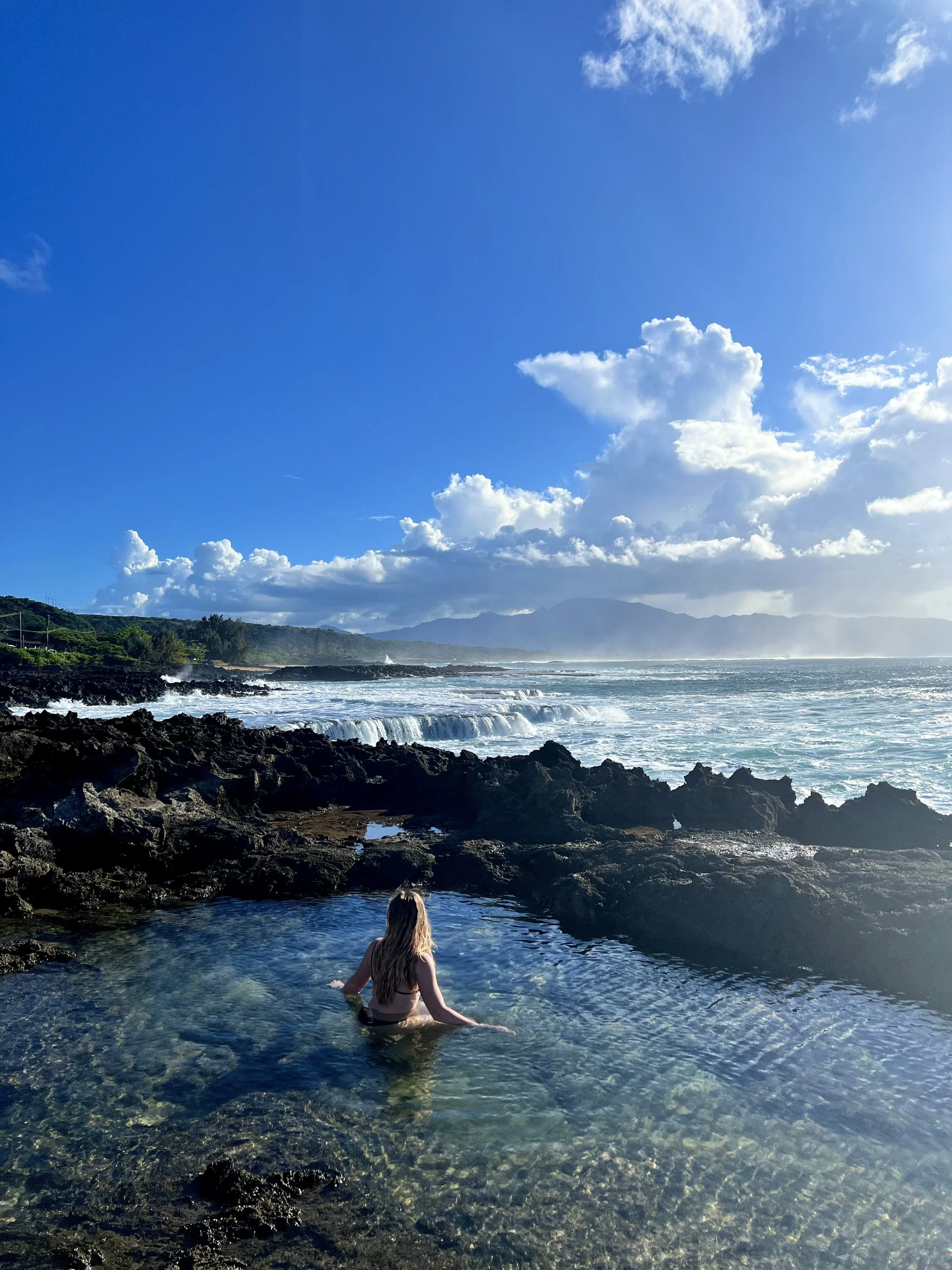 A woman sitting in a natural hot spring by the rocky shoreline with ocean waves, cloudy sky, and distant mountains in the background.