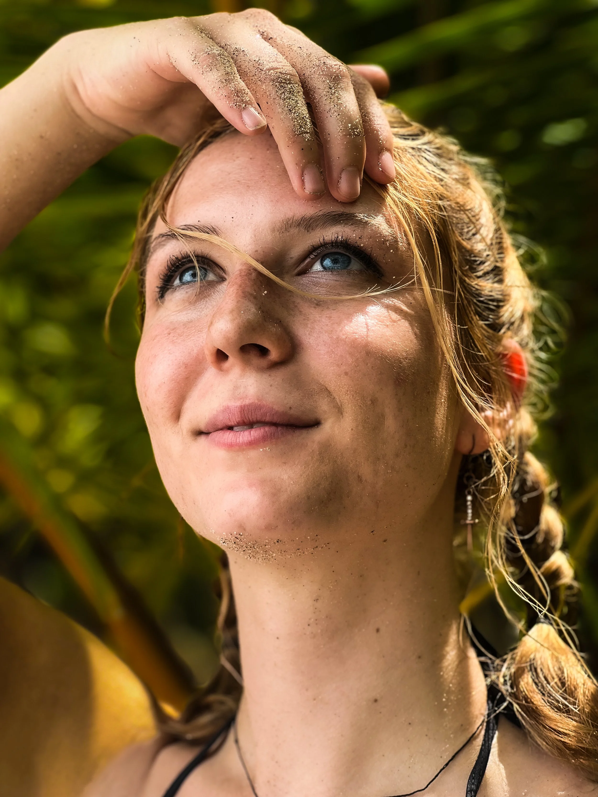 Close-up of a woman with blue eyes and braided hair, looking up, with sunlight on her face, surrounded by green foliage, with sand on her hand and face.