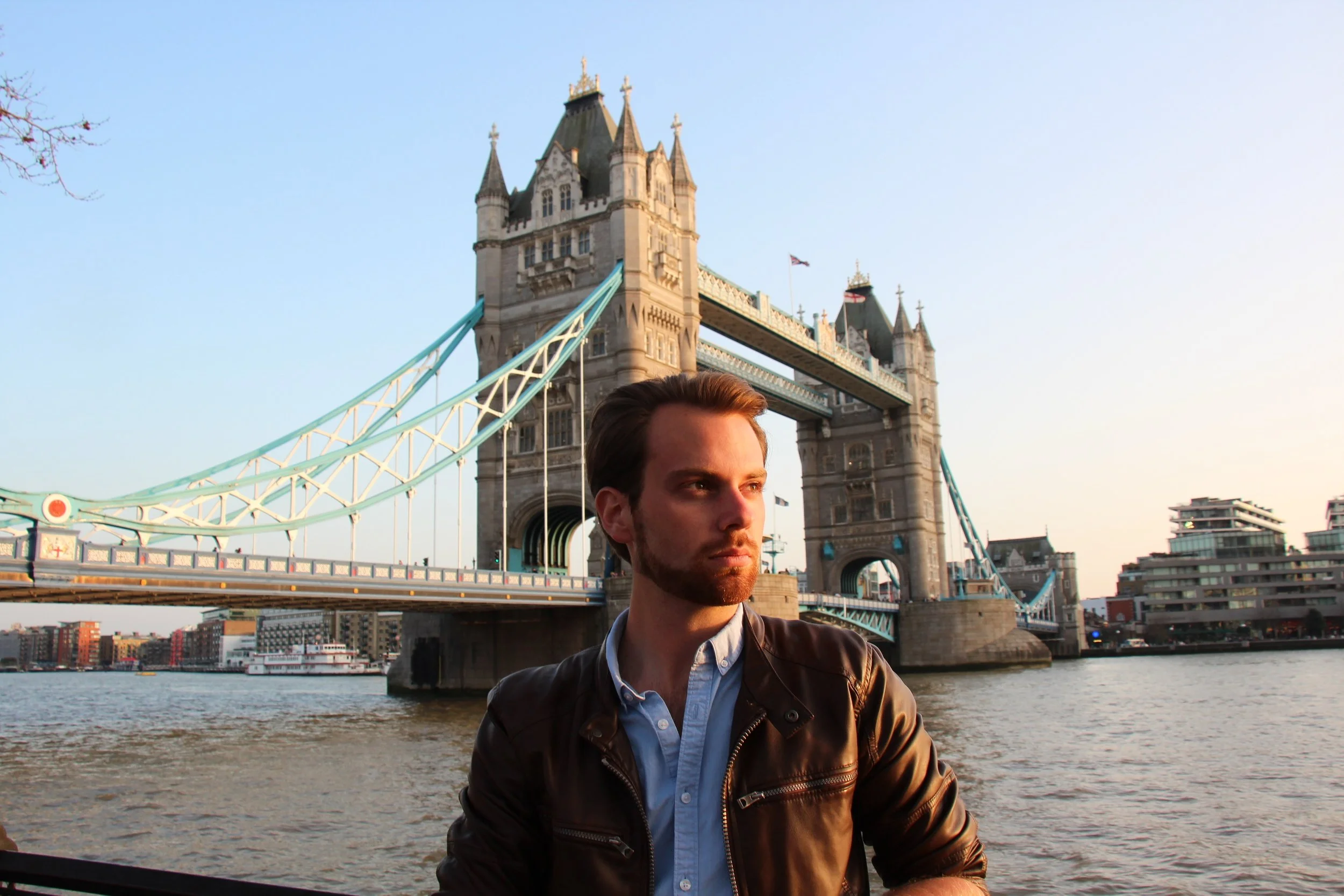 A man with brown hair and a beard, wearing a black leather jacket and light blue shirt, standing near the water with Tower Bridge in London in the background during golden hour sunset.