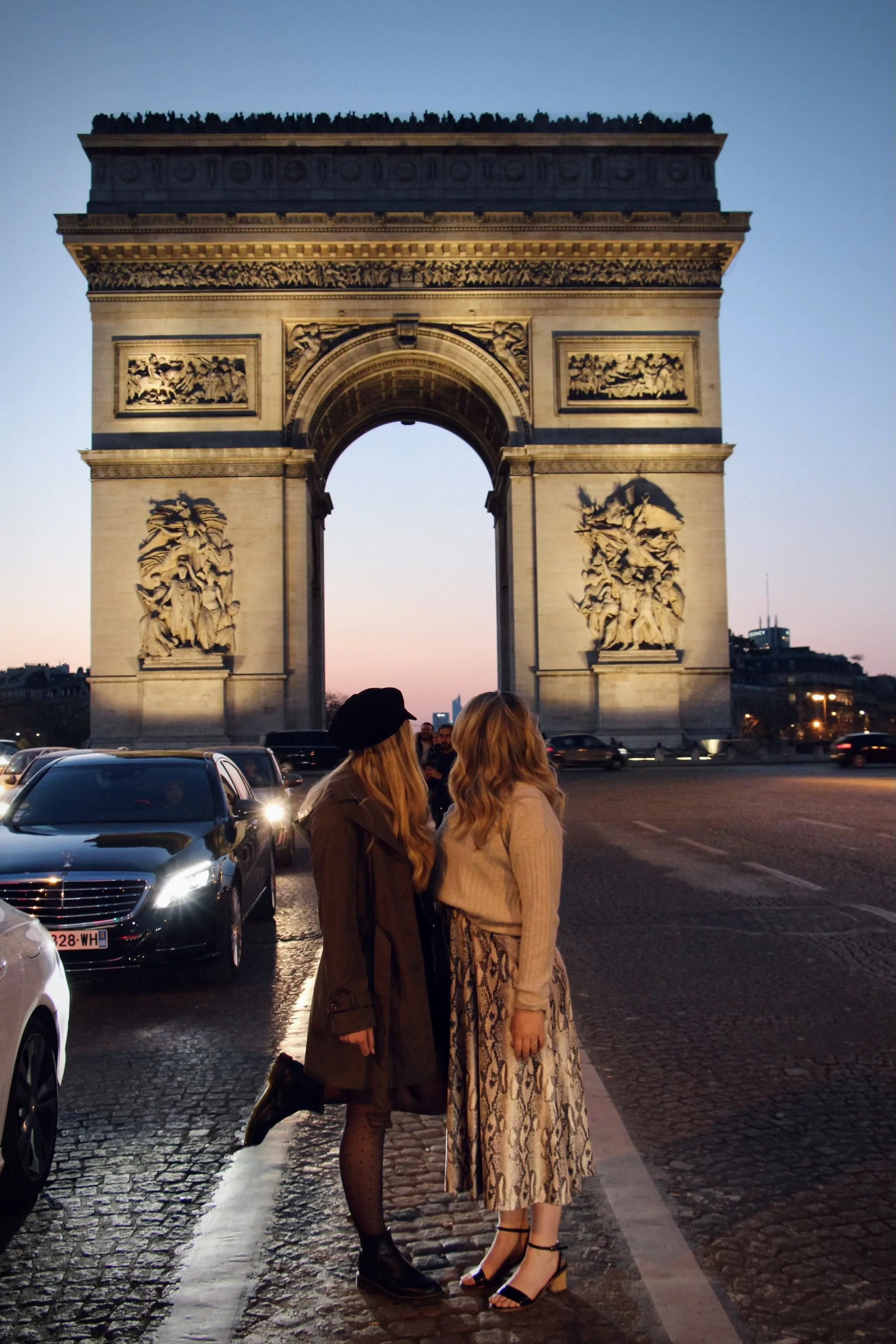 Two women standing in front of the Arc de Triomphe in Paris at dusk, with cars and buildings in the background.