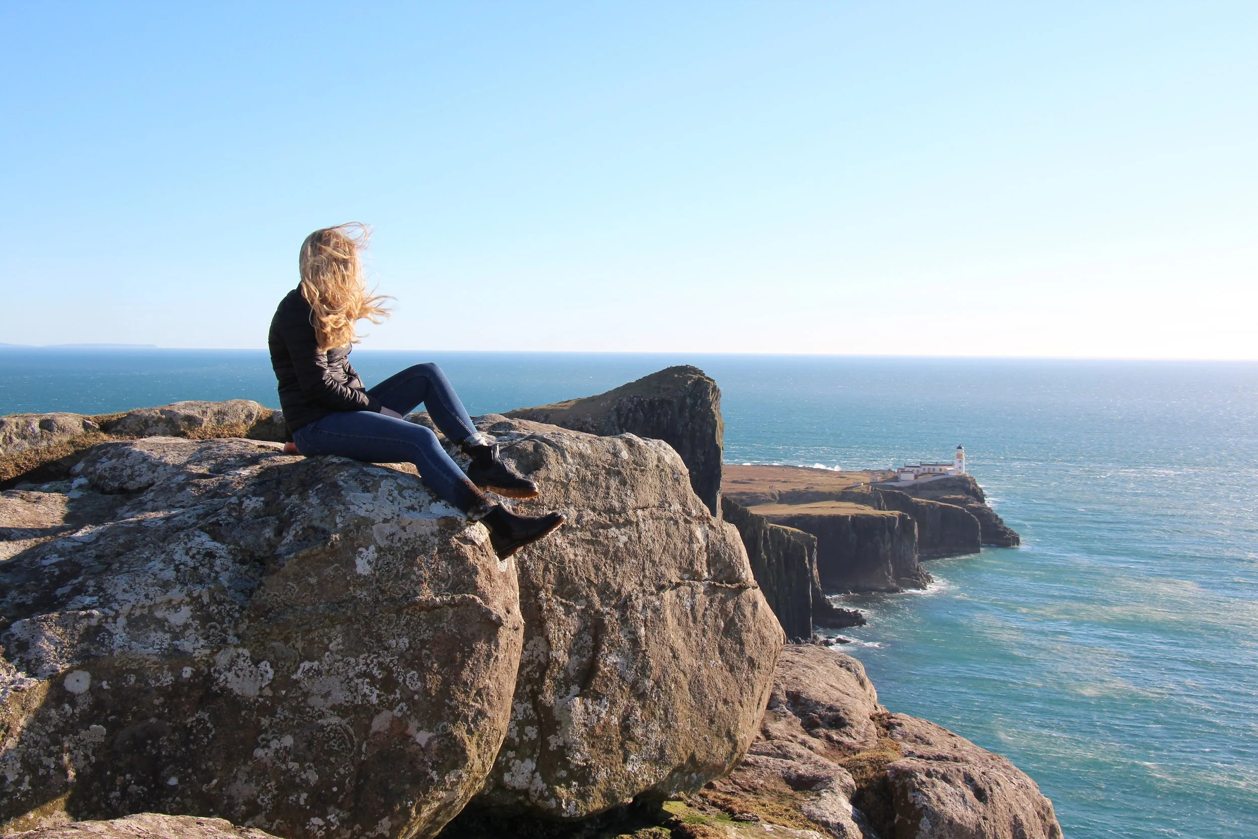 A woman with blonde hair sitting on large rocks overlooking the ocean with cliffs and a lighthouse in the distance, under a clear sky.