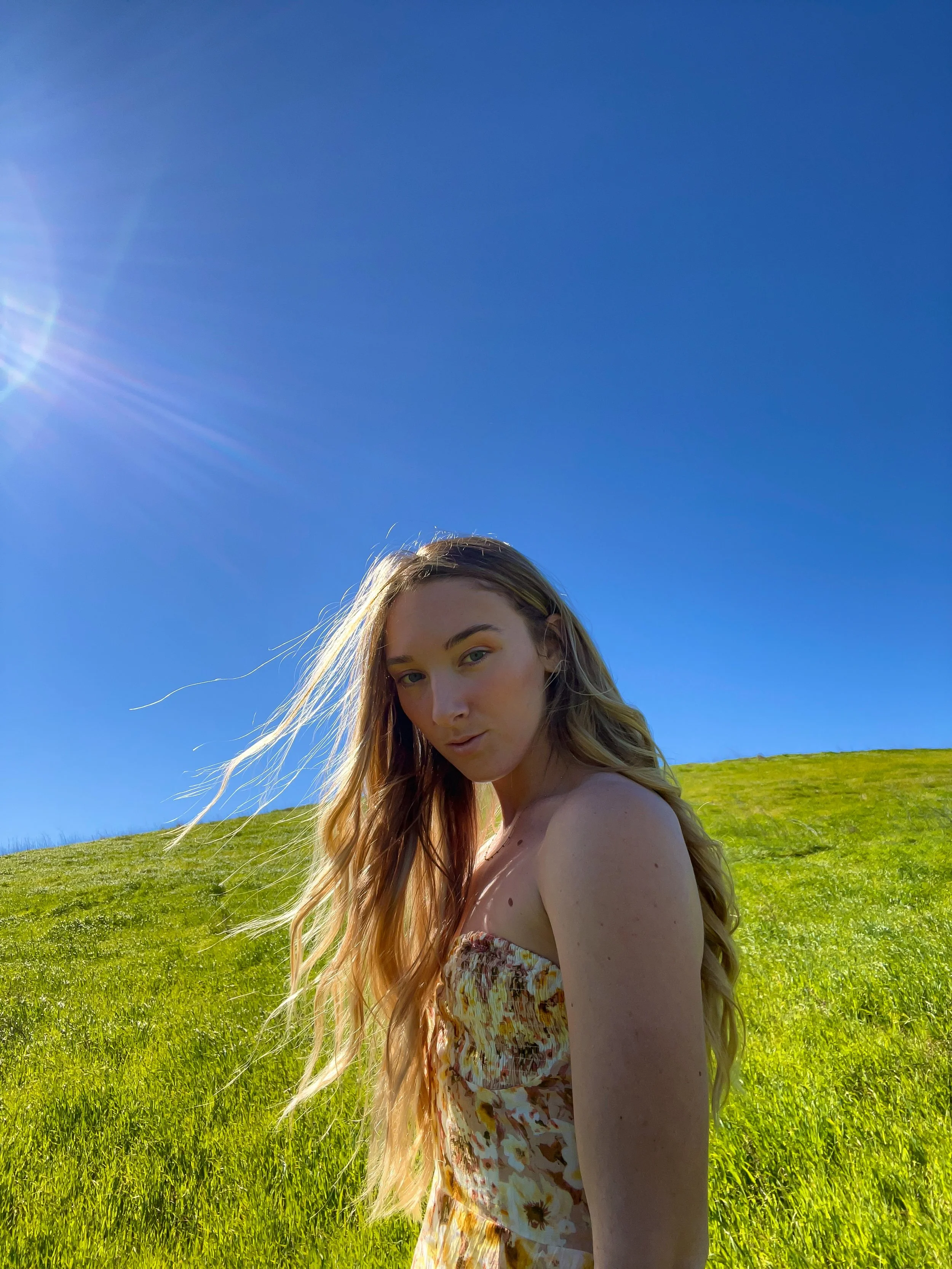 A young woman with long blonde hair standing in a grassy field under a clear blue sky, with the sun shining overhead.