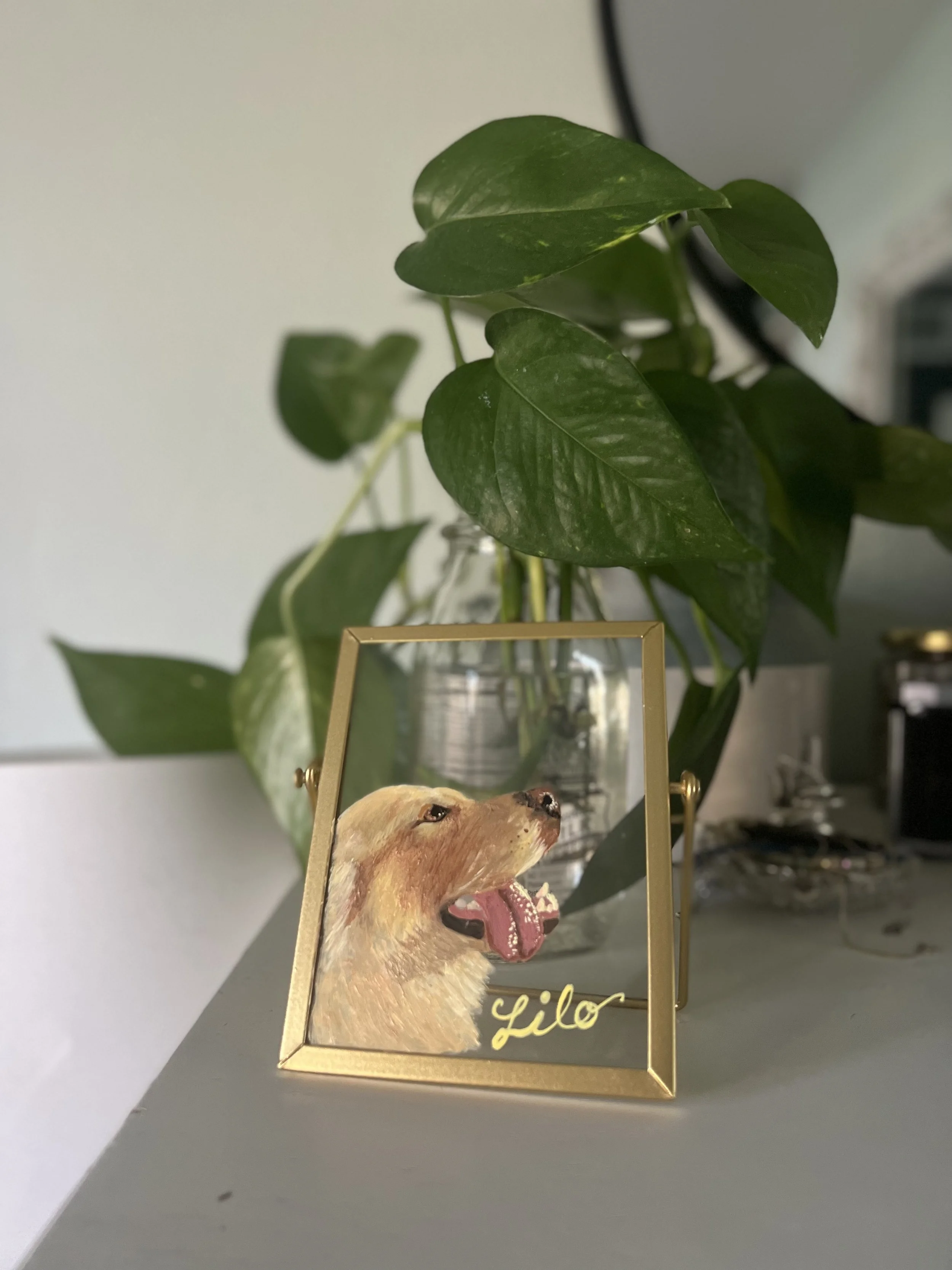 Decorative photo frame with a painting of a yawning golden retriever puppy, with the name "Lilo" written at the bottom, placed on a surface with green leafy plant behind it.