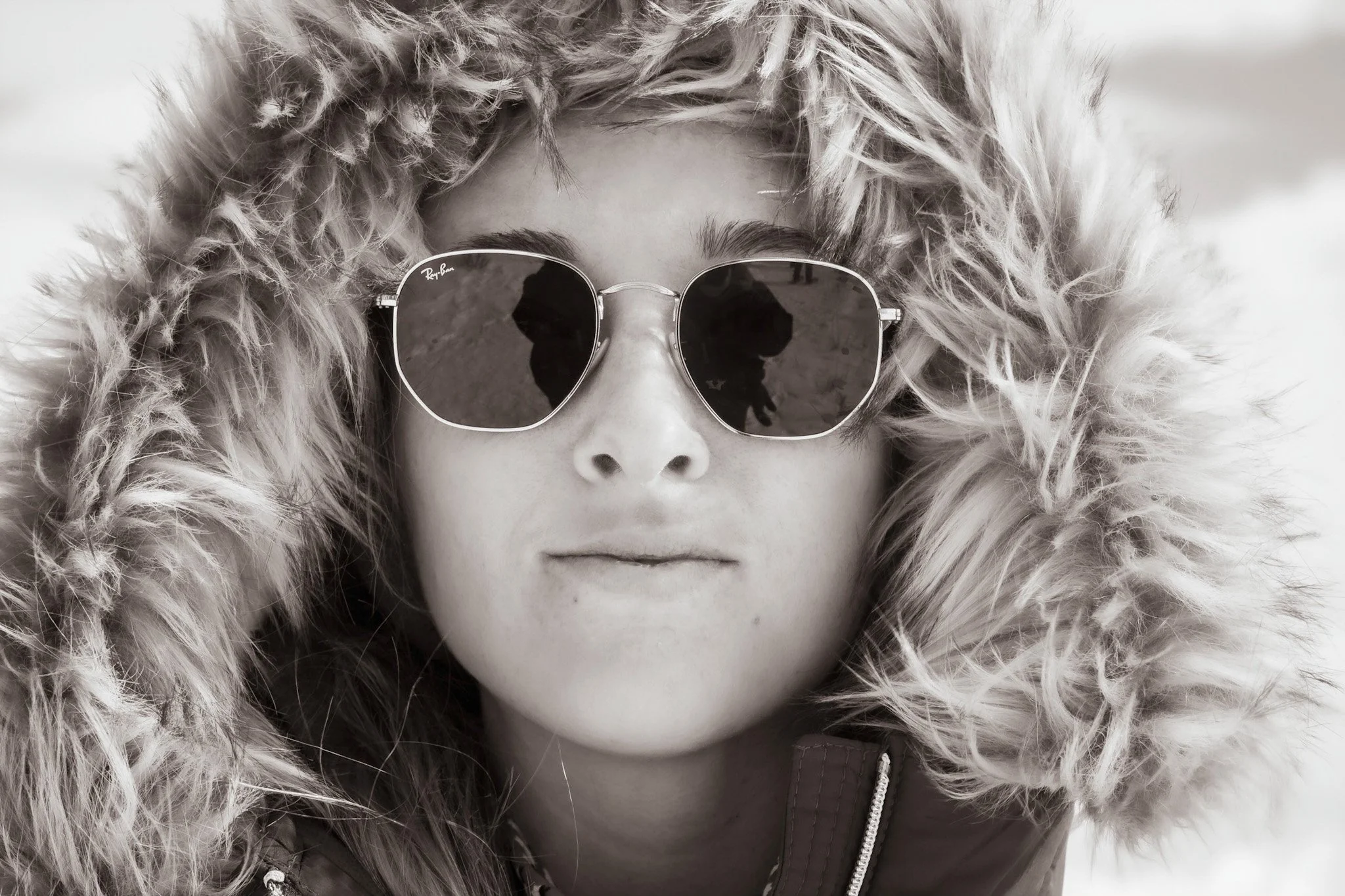 Close-up of a woman with a fur-lined hood wearing sunglasses, with the reflection of a person taking her photo visible in the lenses.