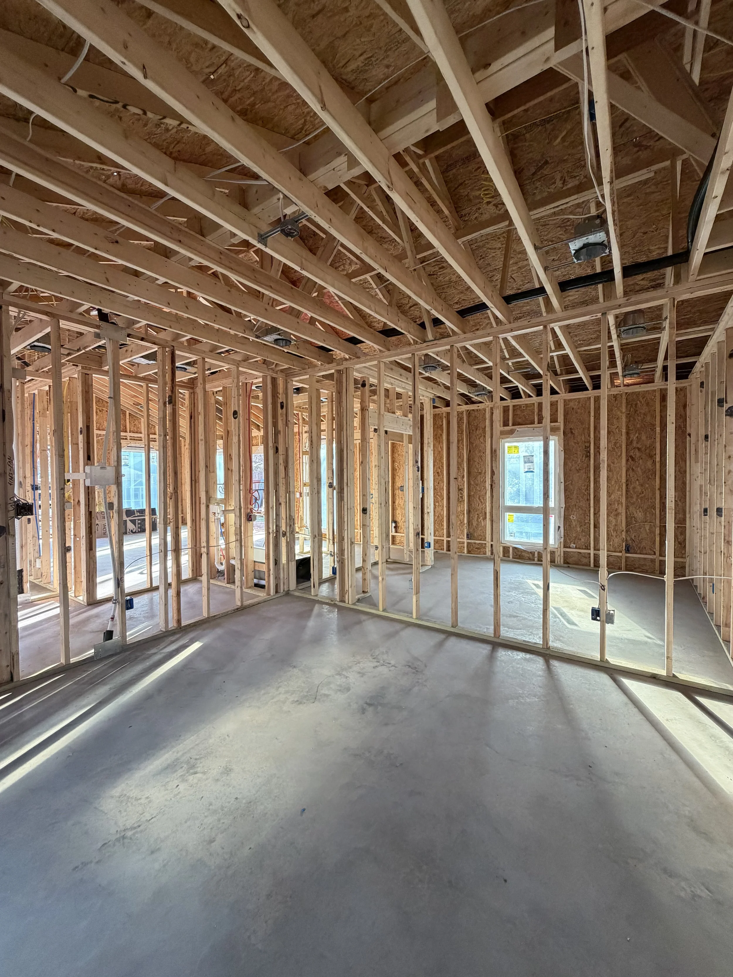 Interior of a house under construction with exposed wooden framing, electrical wiring, and concrete floor, with some windows installed.