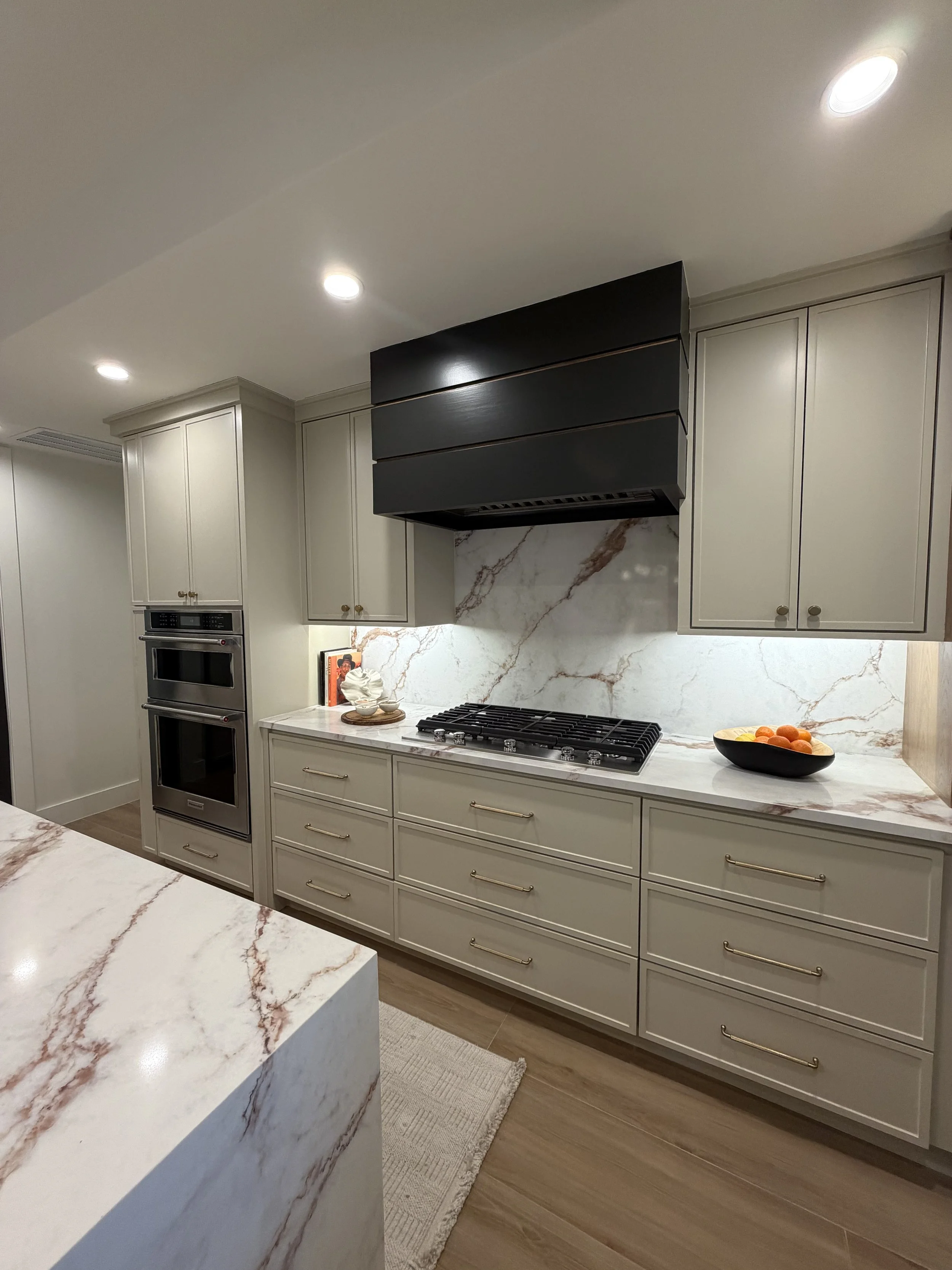Modern kitchen with white cabinetry, marble backsplash, black range hood, built-in oven, and a bowl of oranges on the counter.