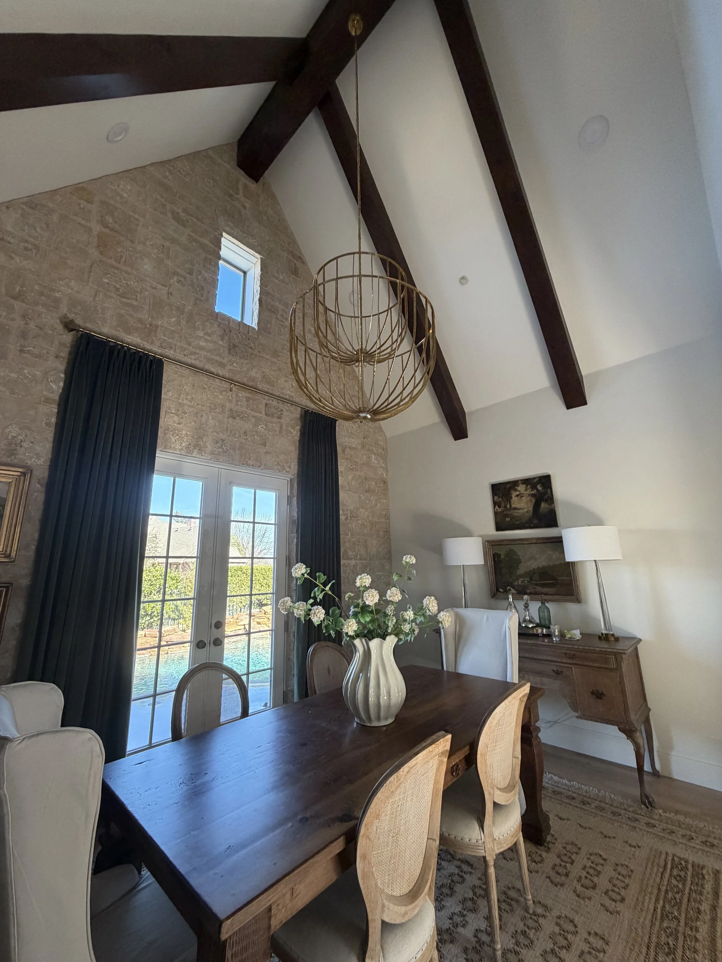 A dining room with a high vaulted ceiling, exposed wooden beams, a chandelier, a stone wall, a large window with navy curtains, a wooden table with a flower vase, and artwork on the wall.