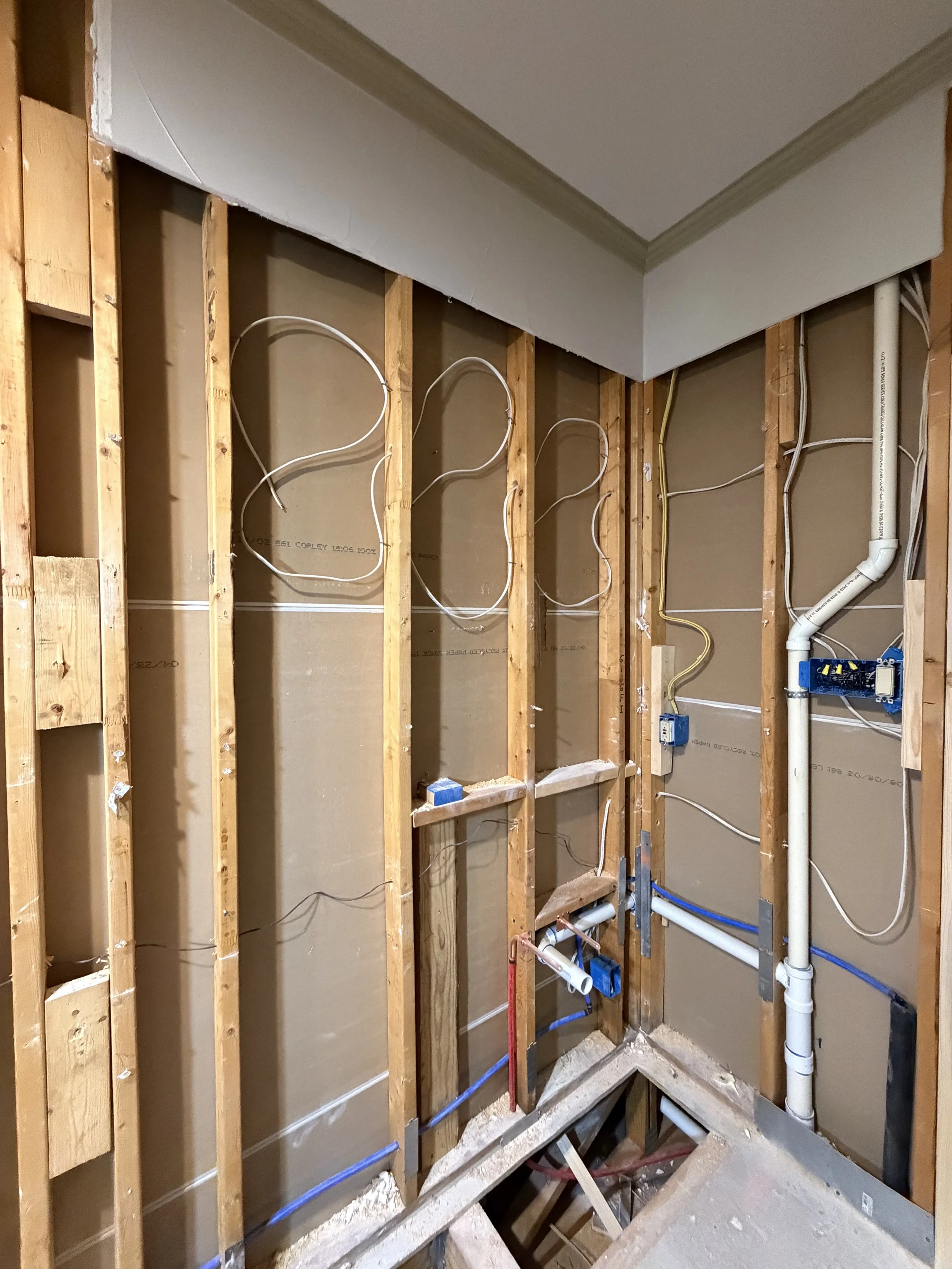 Interior of a room under construction with exposed wooden studs, electrical wiring, and plumbing pipes, with some drywall installed near the ceiling.