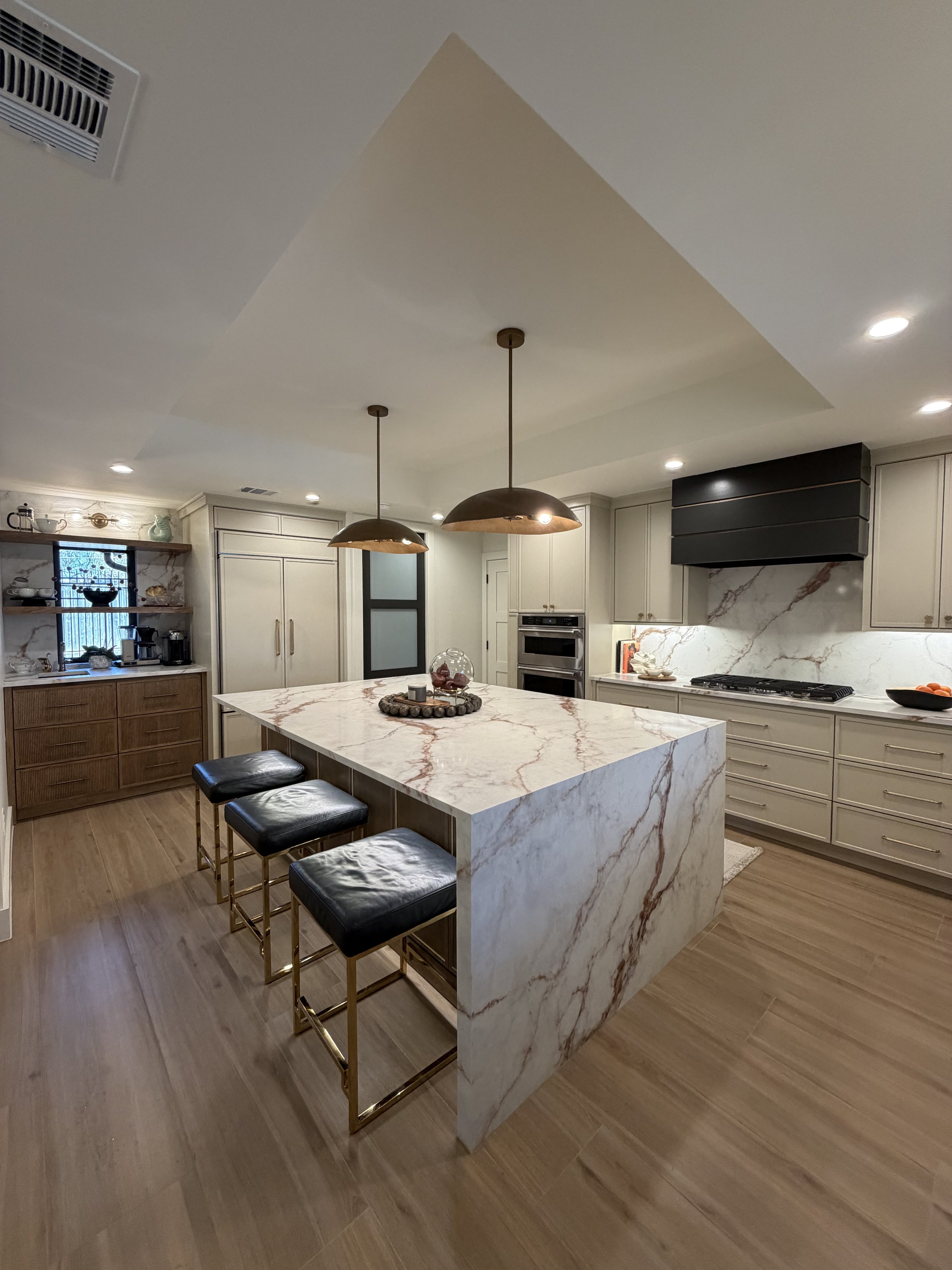 Modern kitchen featuring a marble island with three black and gold barstools, white cabinetry, built-in double oven, black range hood, wooden floor, and pendant lighting.