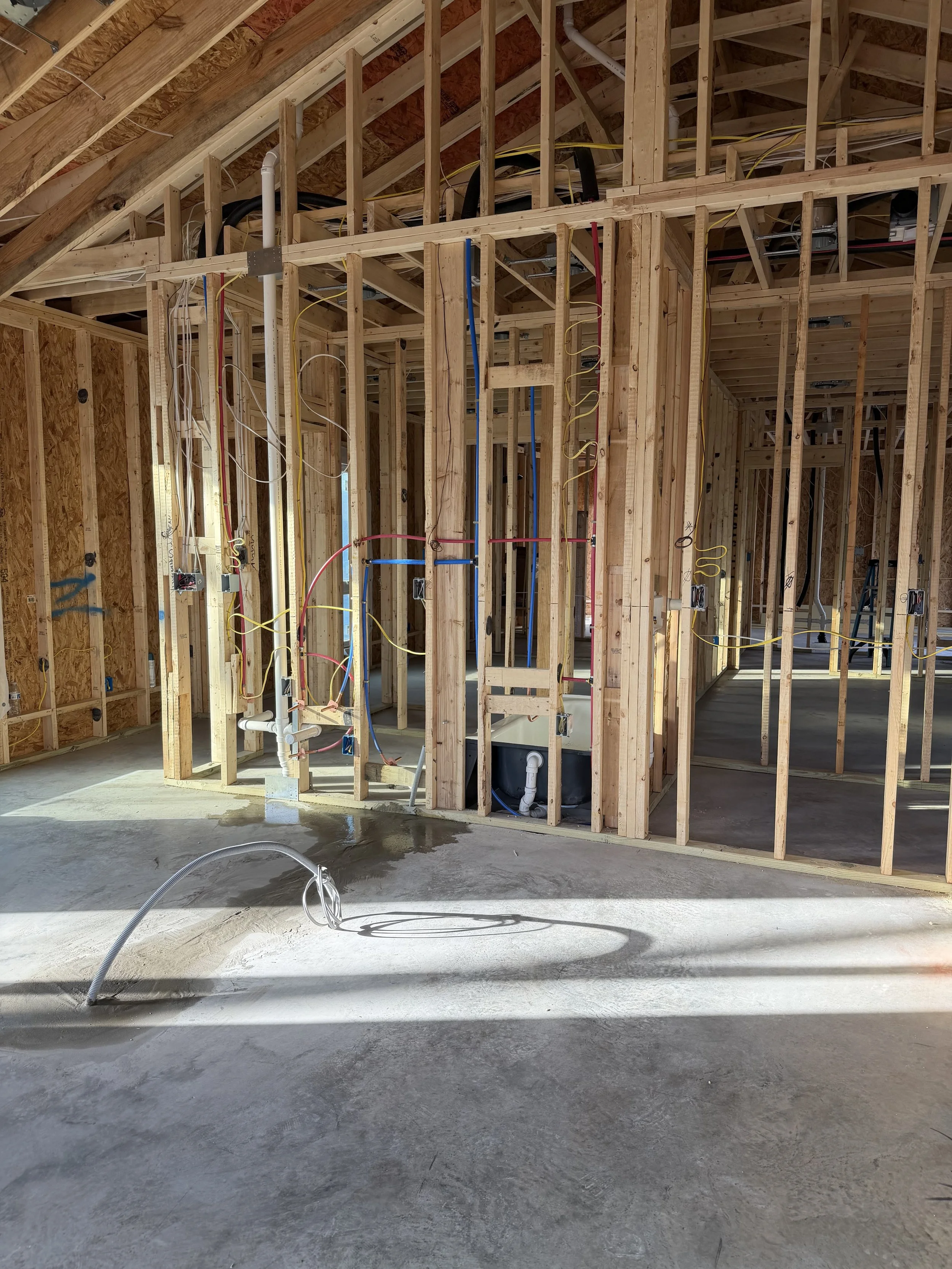 Construction site of a house with exposed wooden framing, electrical wiring, plumbing pipes, and a concrete floor with water on it.