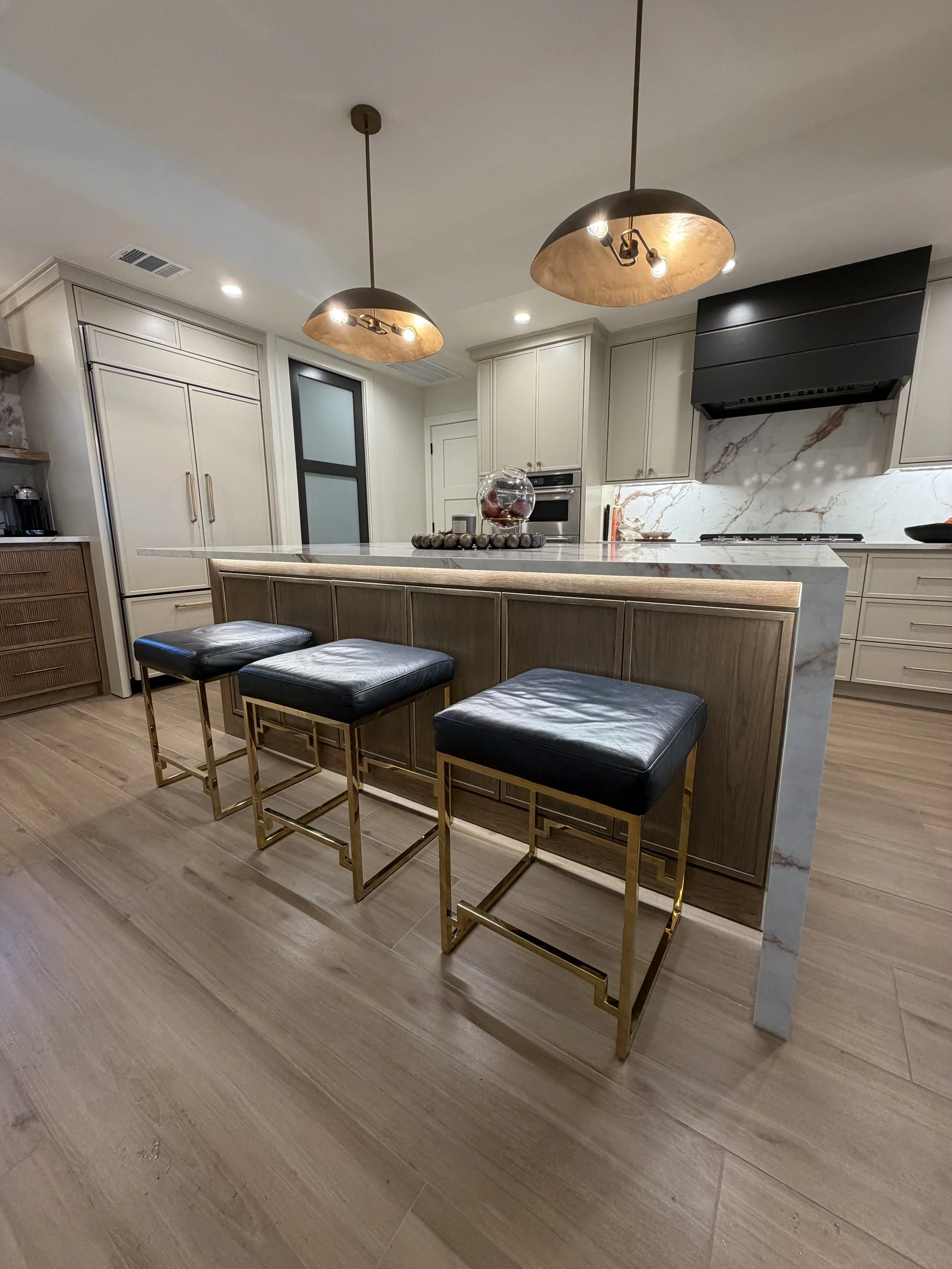 Modern kitchen with a large island, three black and gold bar stools, pendant lights, and white cabinetry with marble backsplash.