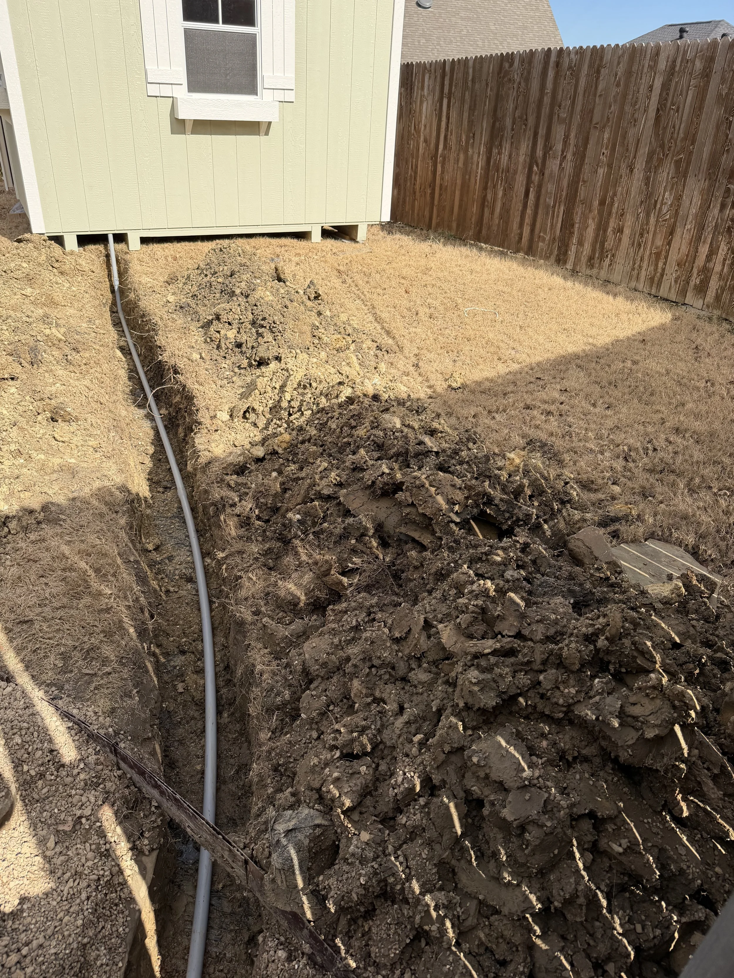 A backyard with a trench dug for laying pipes or cables, next to a house, with a pile of dirt and a wooden fence in the background.