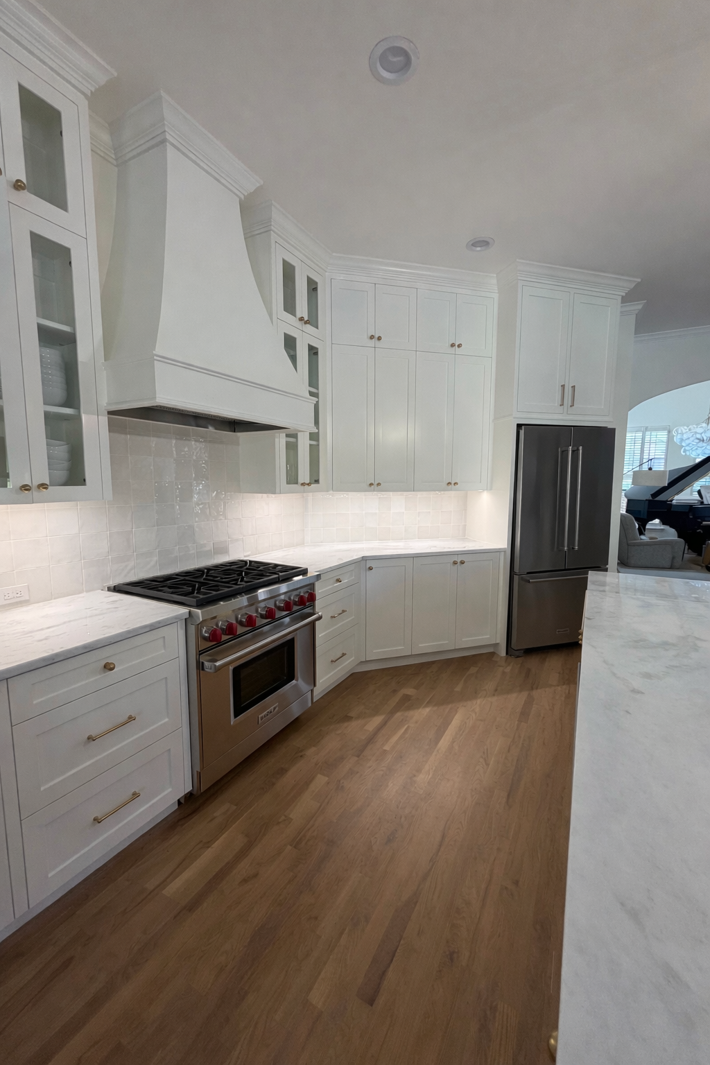 Modern kitchen with white cabinets, marble countertops, and stainless steel appliances including a refrigerator and oven. Hardwood floors and a white tiled backsplash.