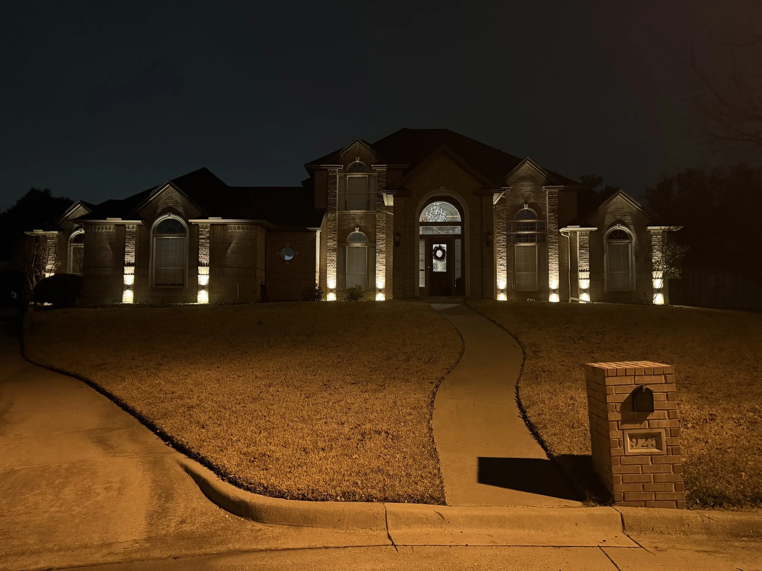 A large brick house at night with landscape lighting illuminating the front exterior, a curved concrete walkway leading up to the front door, and a brick mailbox with house number 9245 in the front yard.