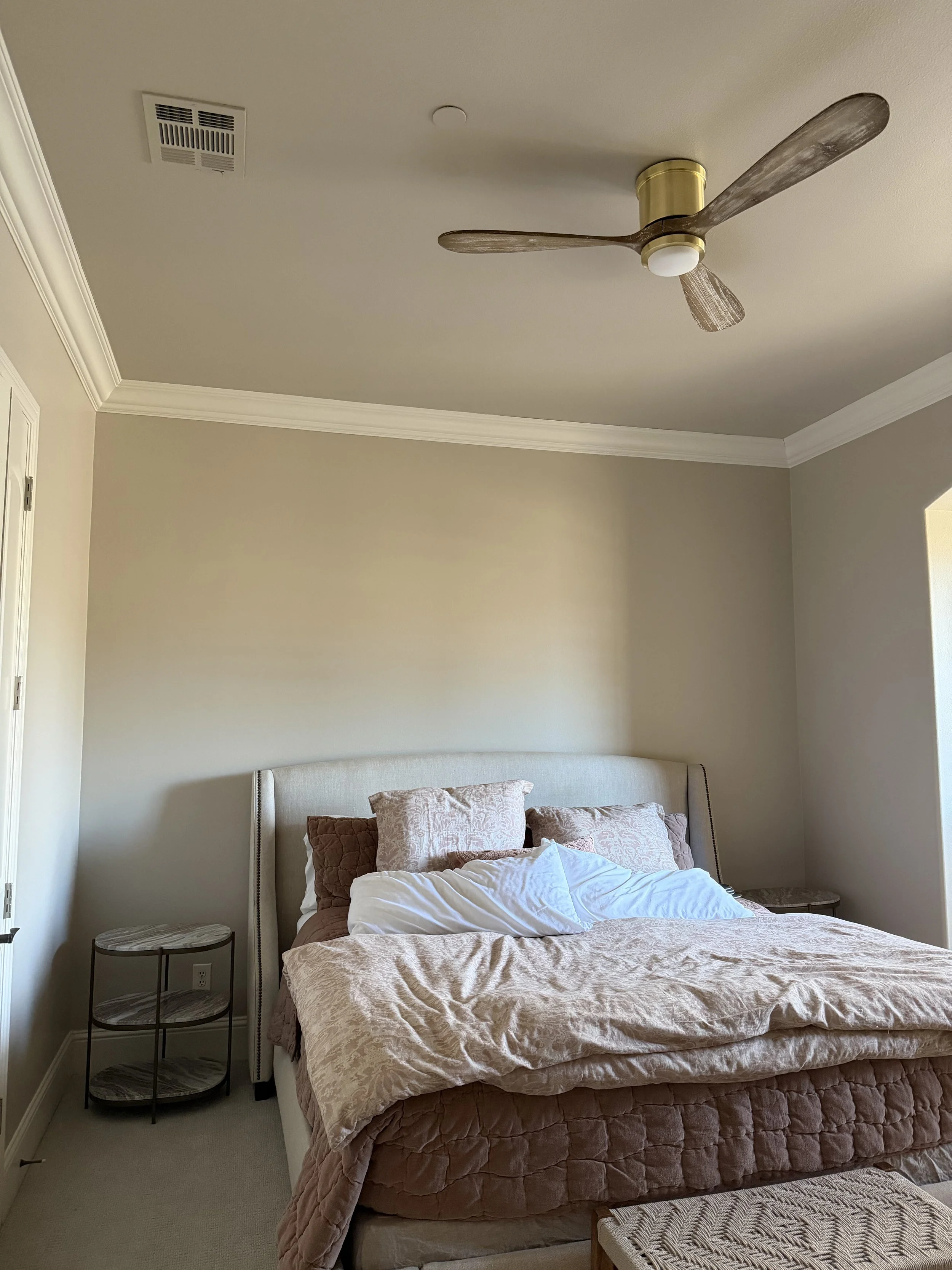 A bedroom with a beige headboard, thrown blanket, multiple pillows, two round side tables, a ceiling fan, and a vent on the ceiling.