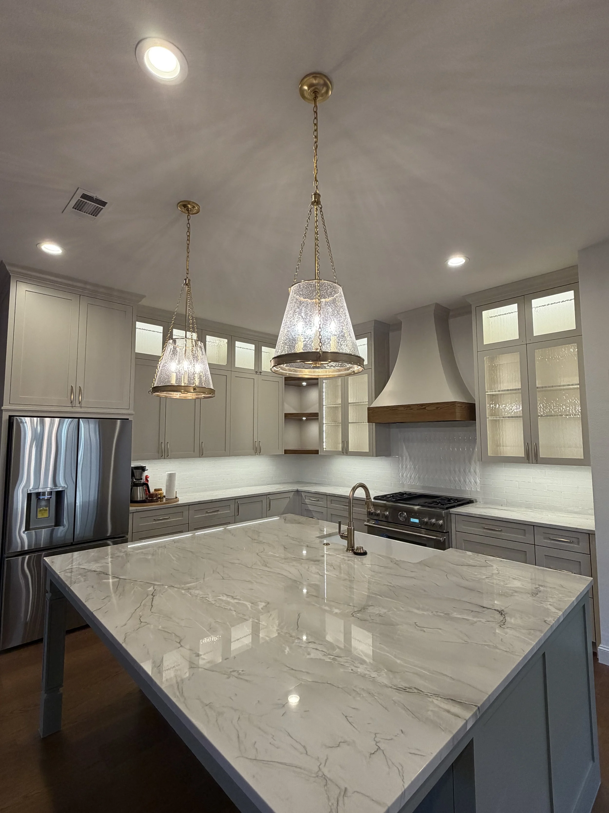 Modern kitchen with white marble island, gray cabinetry, stainless steel refrigerator, and two hanging pendant lights.
