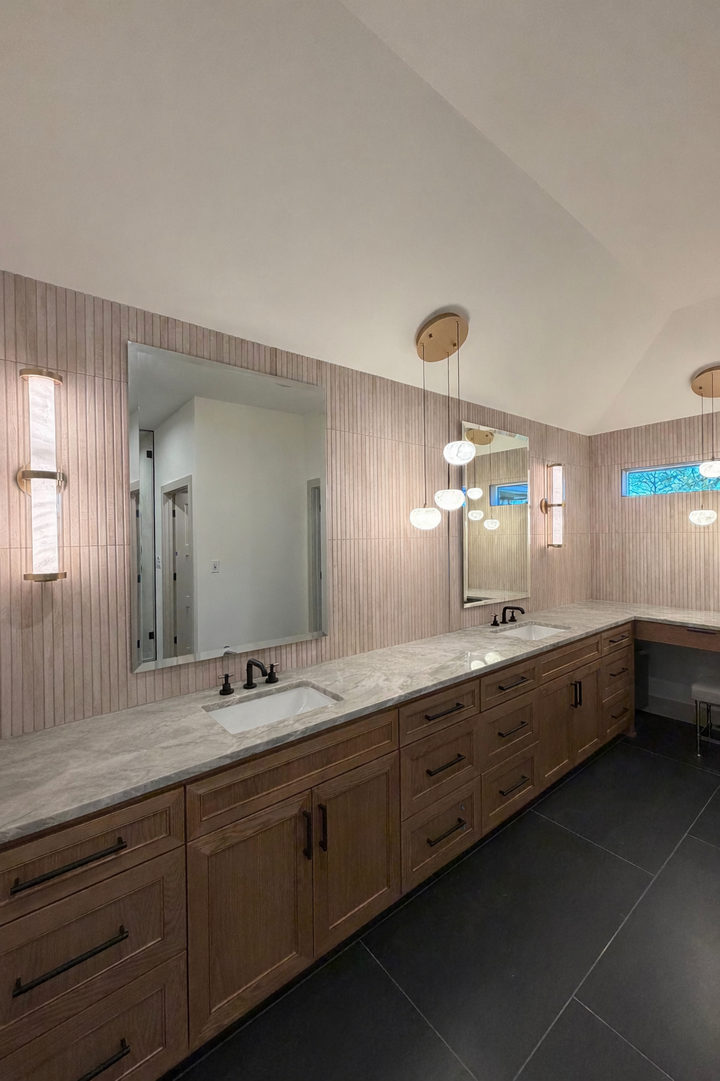 Modern bathroom vanity with dual sinks, marble countertop, wooden cabinets, wall-mounted mirrors, pendant and wall lighting, small window with tree view.