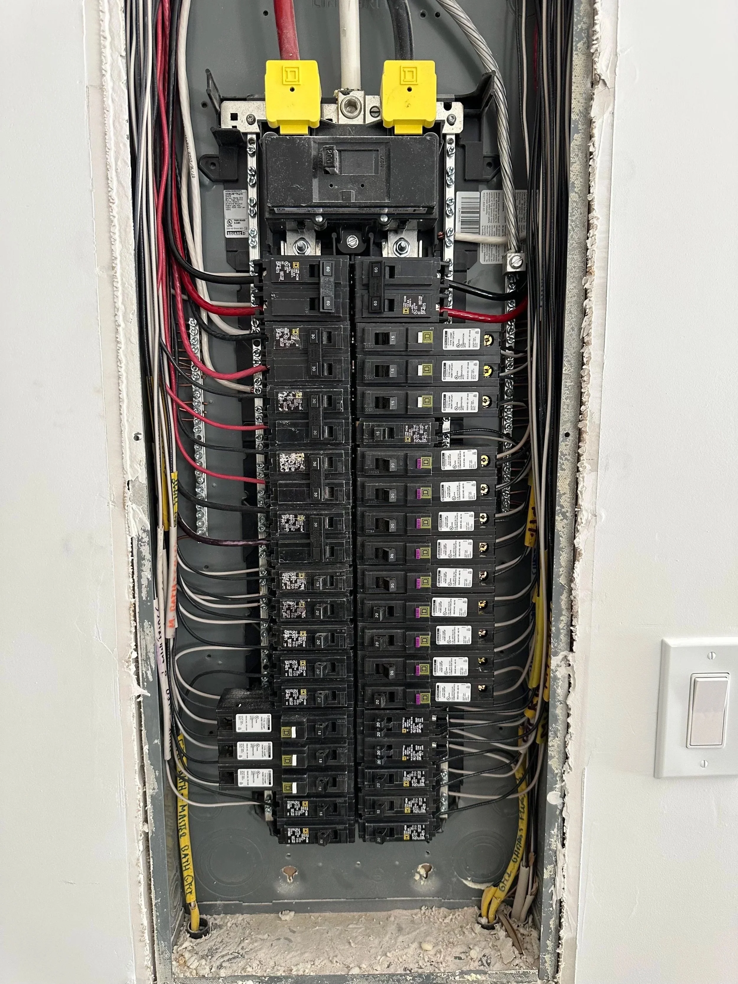 Electrical breaker panel with multiple circuit breakers, wiring, and yellow warning labels, partially installed in a wall with exposed drywall.