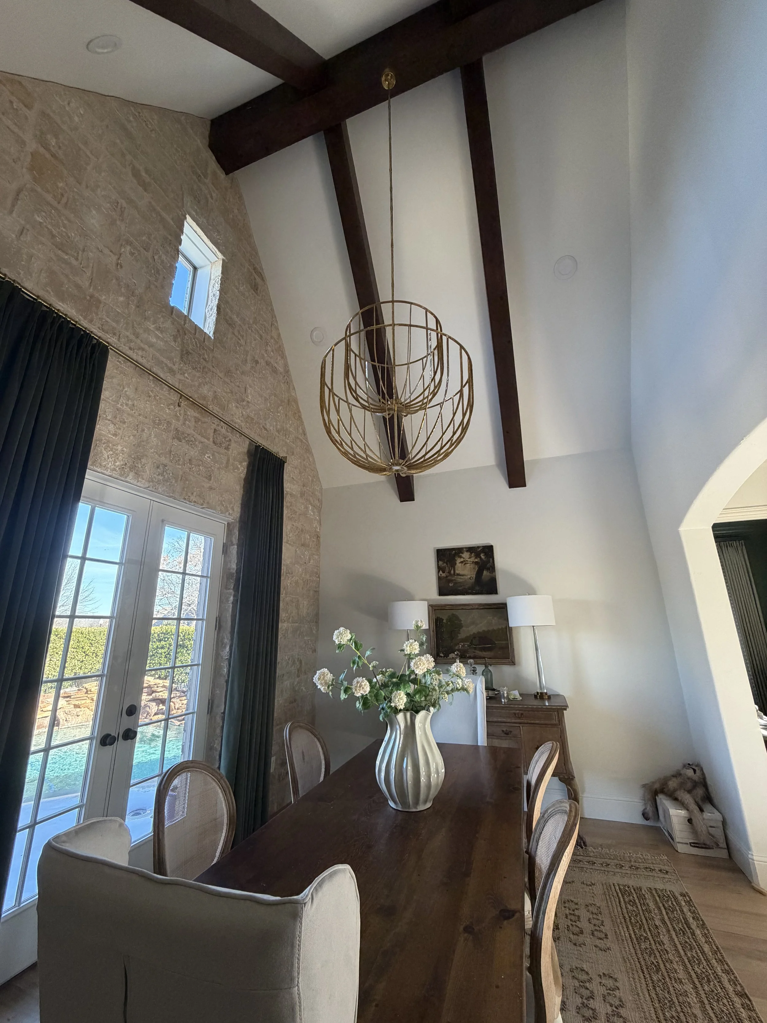 A dining room with a high vaulted ceiling, exposed wooden beams, and a large modern chandelier. There are French doors with dark curtains, a wooden dining table with a white vase of flowers, and a sideboard with lamps and artwork. A small window is n