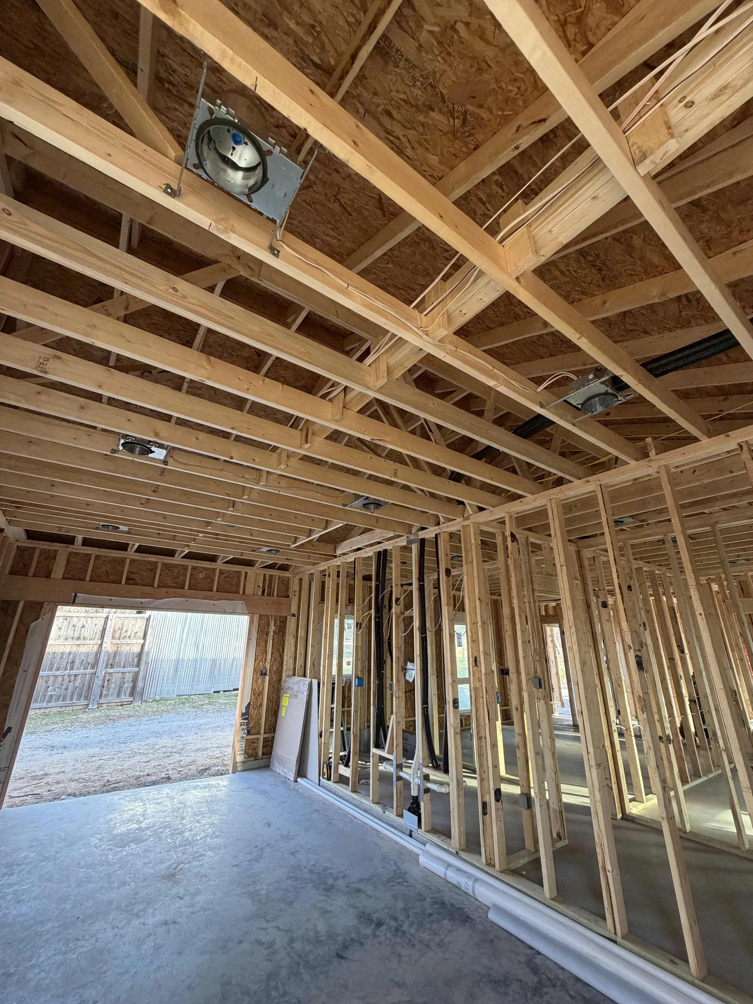 Interior of a house under construction with exposed wooden framing, electrical wiring, and plumbing. The ceiling has wooden beams and metal fixtures for lighting or vents. There is a large open doorway leading outside to a yard.