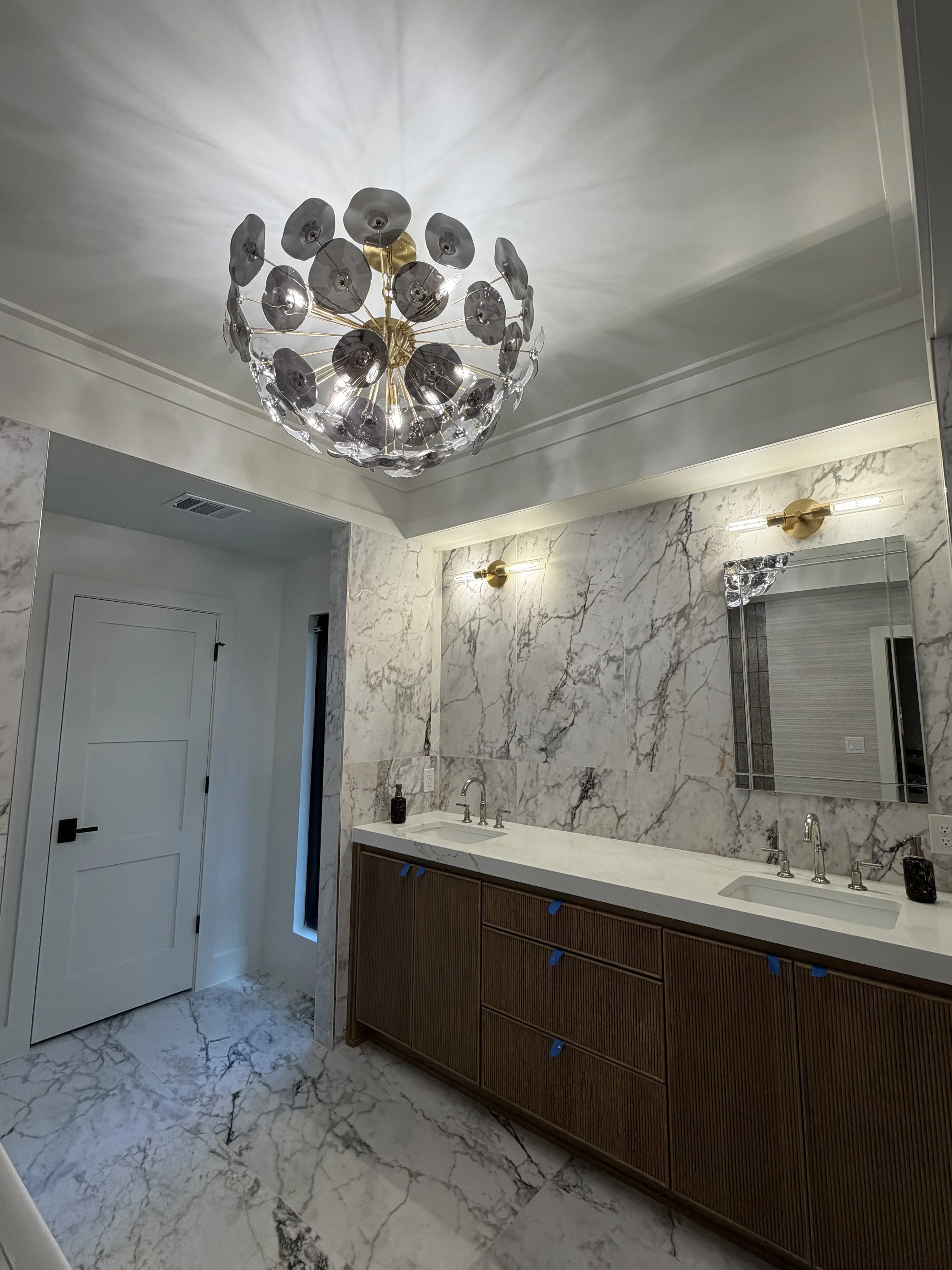 Modern bathroom with marble walls and floors, double sink vanity with wooden cabinetry, large mirror, and decorative lighting fixtures, including a chandelier hanging from the ceiling.
