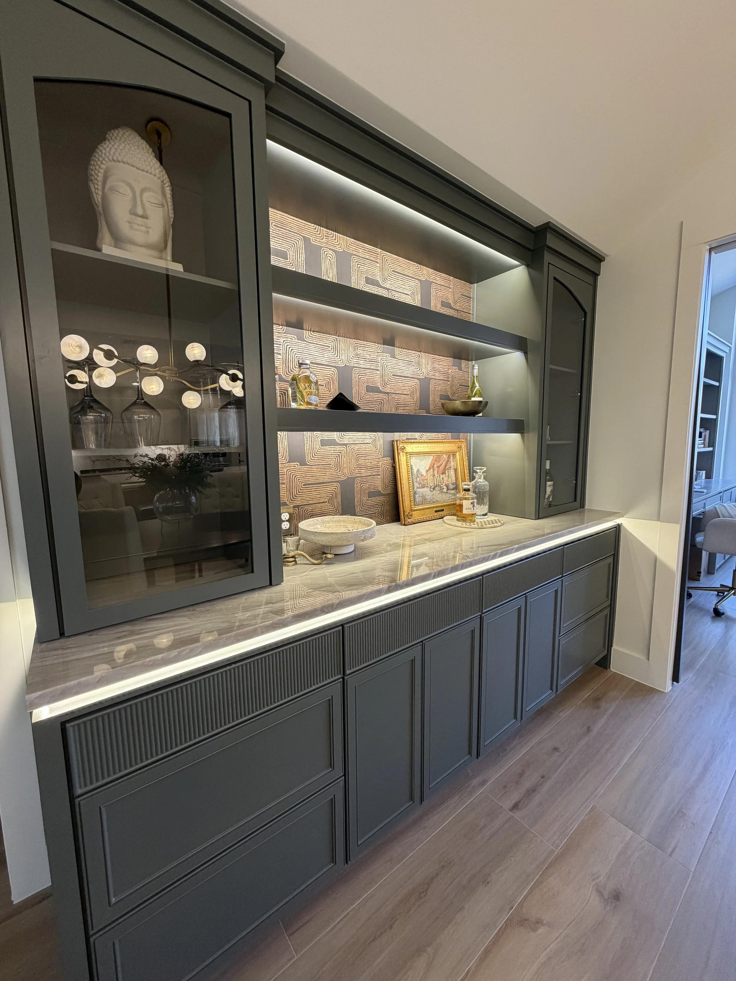 A gray built-in cabinet with a marble countertop, decorated with artworks and glass bottles, featuring a Buddha head sculpture inside a glass cabinet on the left side. There is a decorative wallpaper with geometric pattern behind the open shelves.