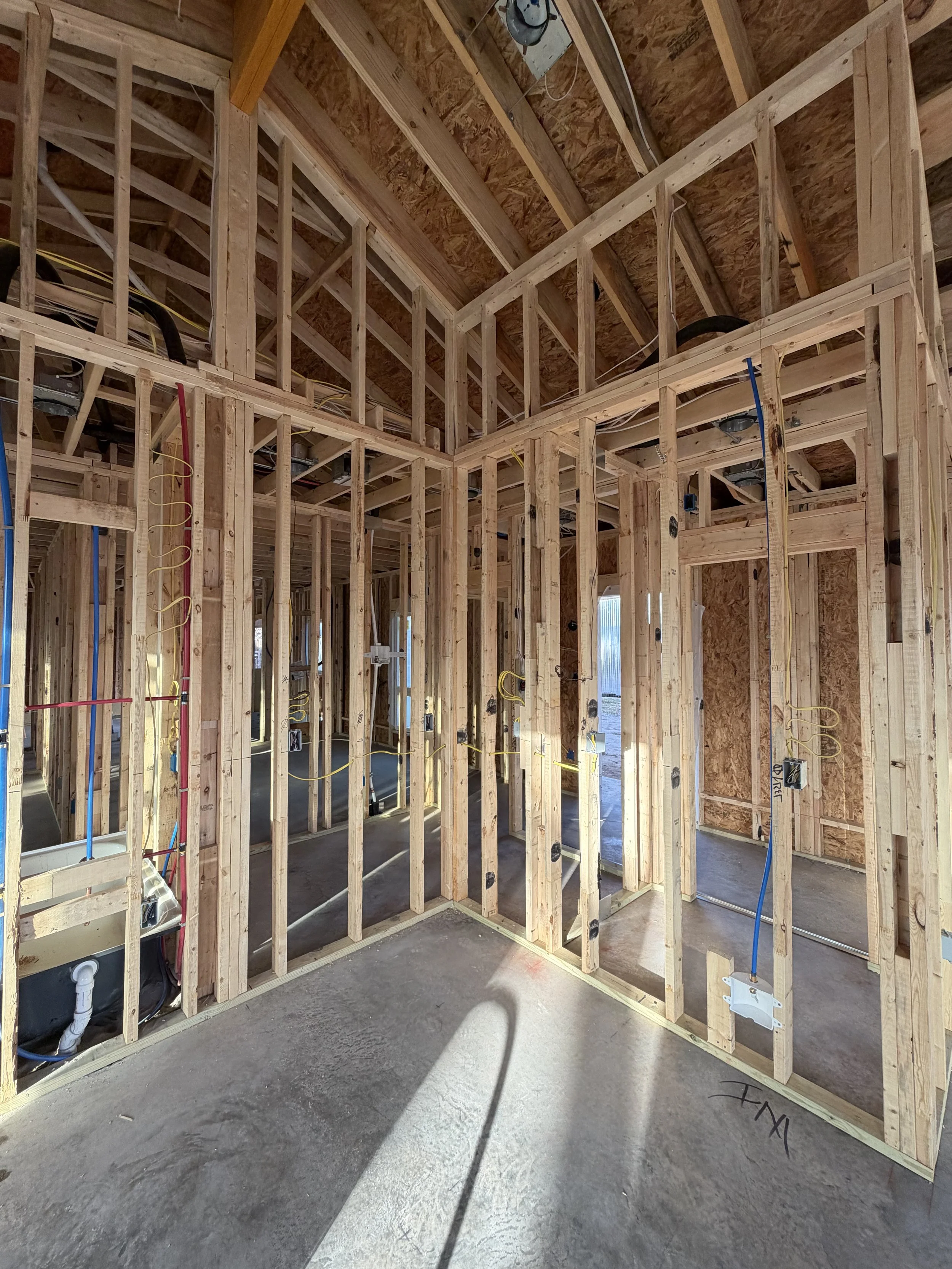 Interior view of a house under construction, showing wooden framing, electrical wiring, and exposed ceiling.