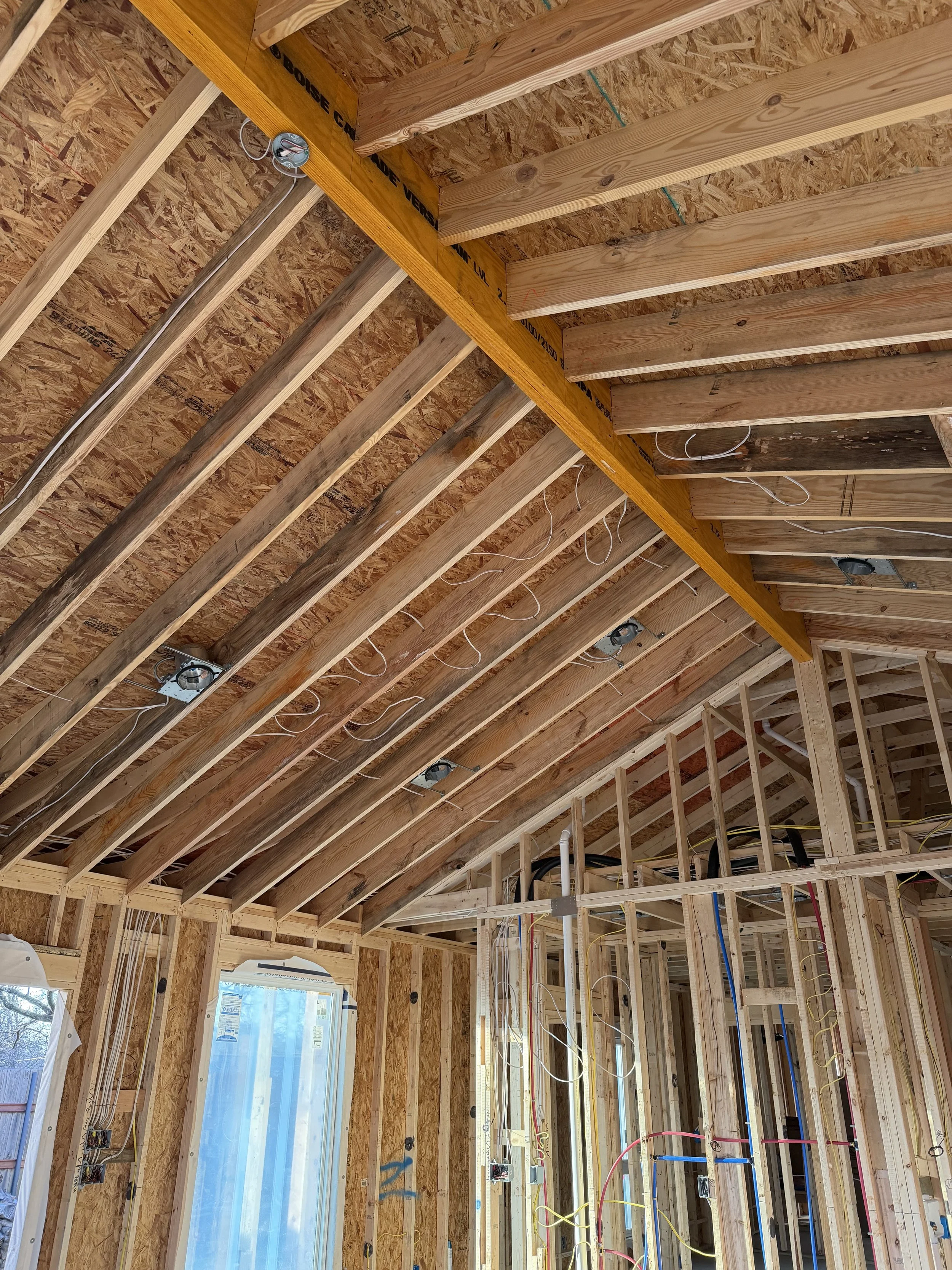 Interior view of a house under construction showing exposed wood framing, electrical wiring, and a ceiling with wooden beams.