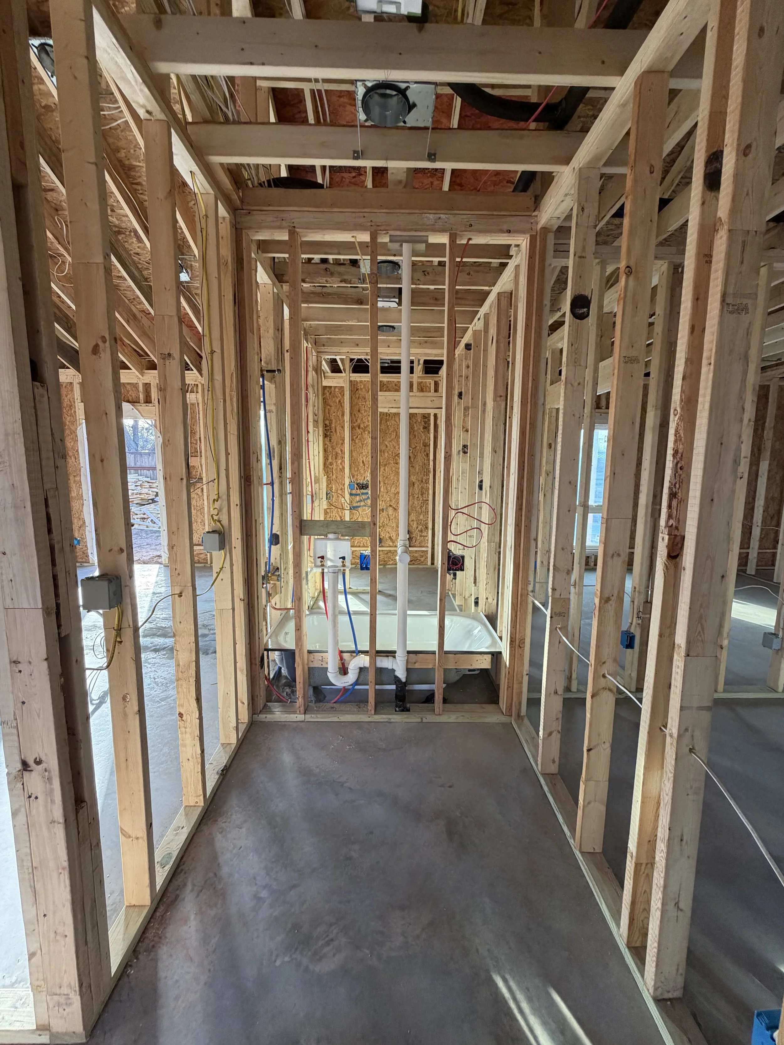 Interior view of a house under construction, showing wooden framing, electrical wiring, plumbing, and a small bathtub in a bathroom area.