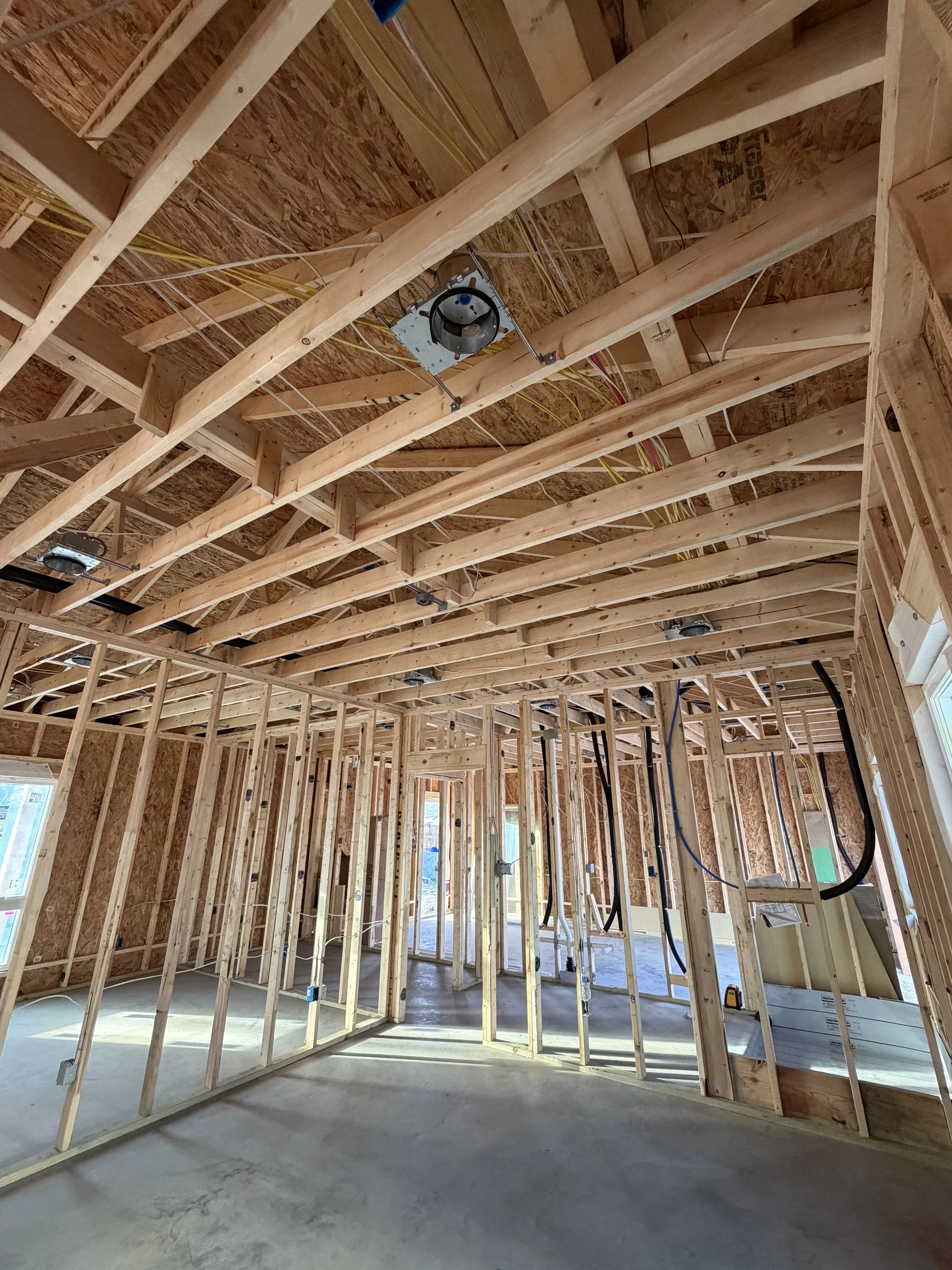 Interior framing of a house under construction with wooden studs, exposed ceiling rafters, wiring, and HVAC ductwork.