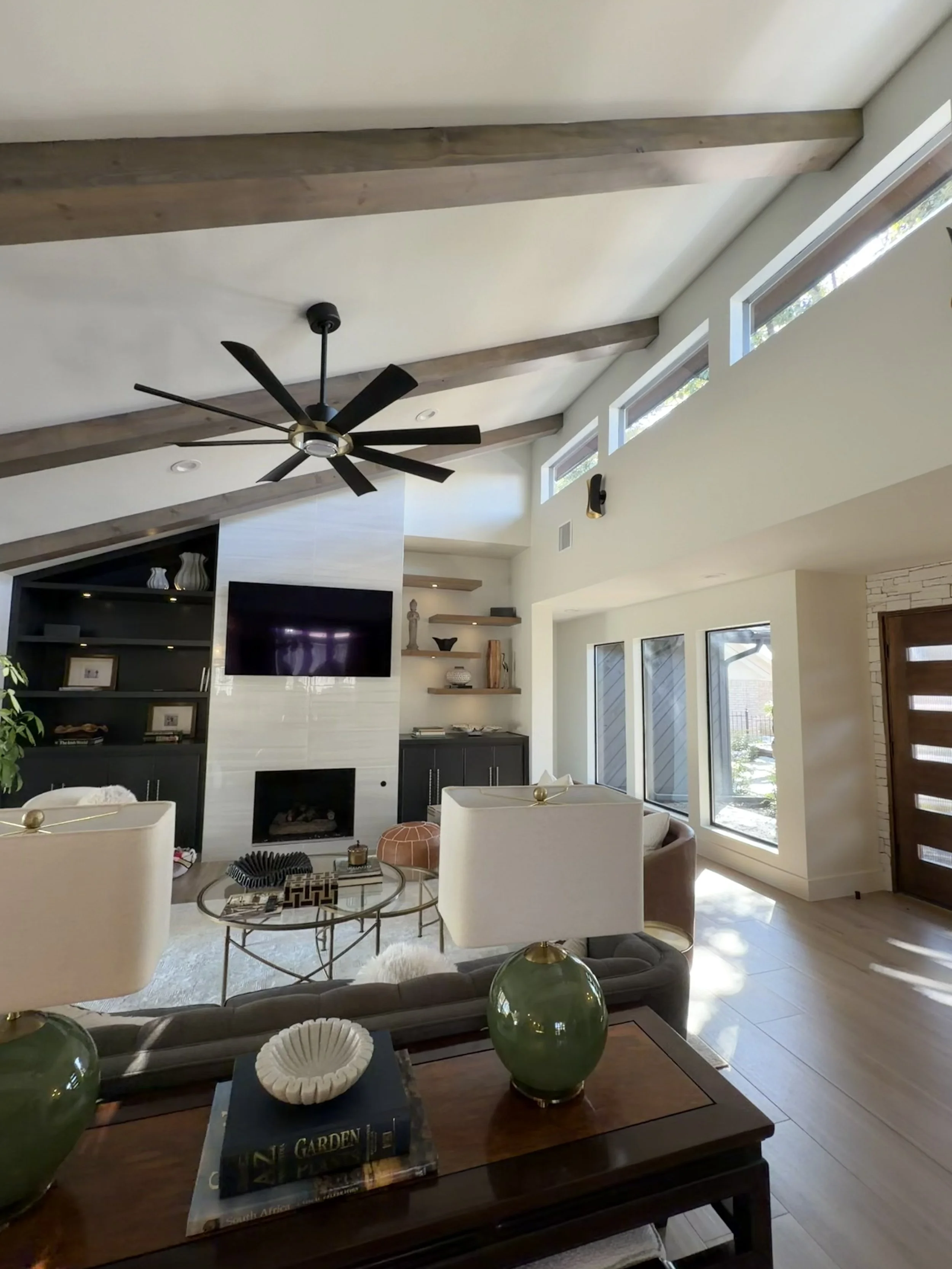 Living room with vaulted ceiling, exposed wooden beams, ceiling fan, large windows, and modern decor.