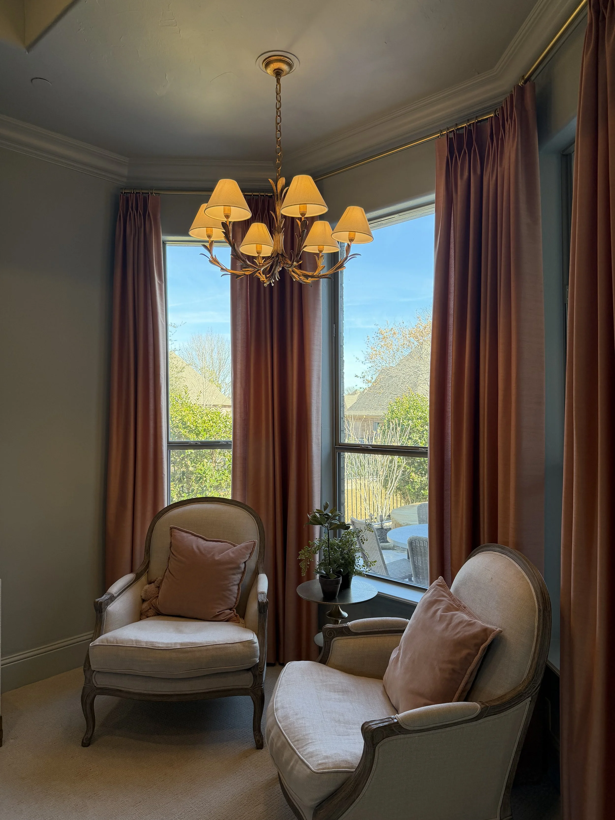 A cozy sitting area with two vintage armchairs with beige upholstery and pink pillows, a small round table with a potted plant, large windows with pink curtains, and a chandelier with multiple small lampshades hanging from the ceiling, overlooking a 