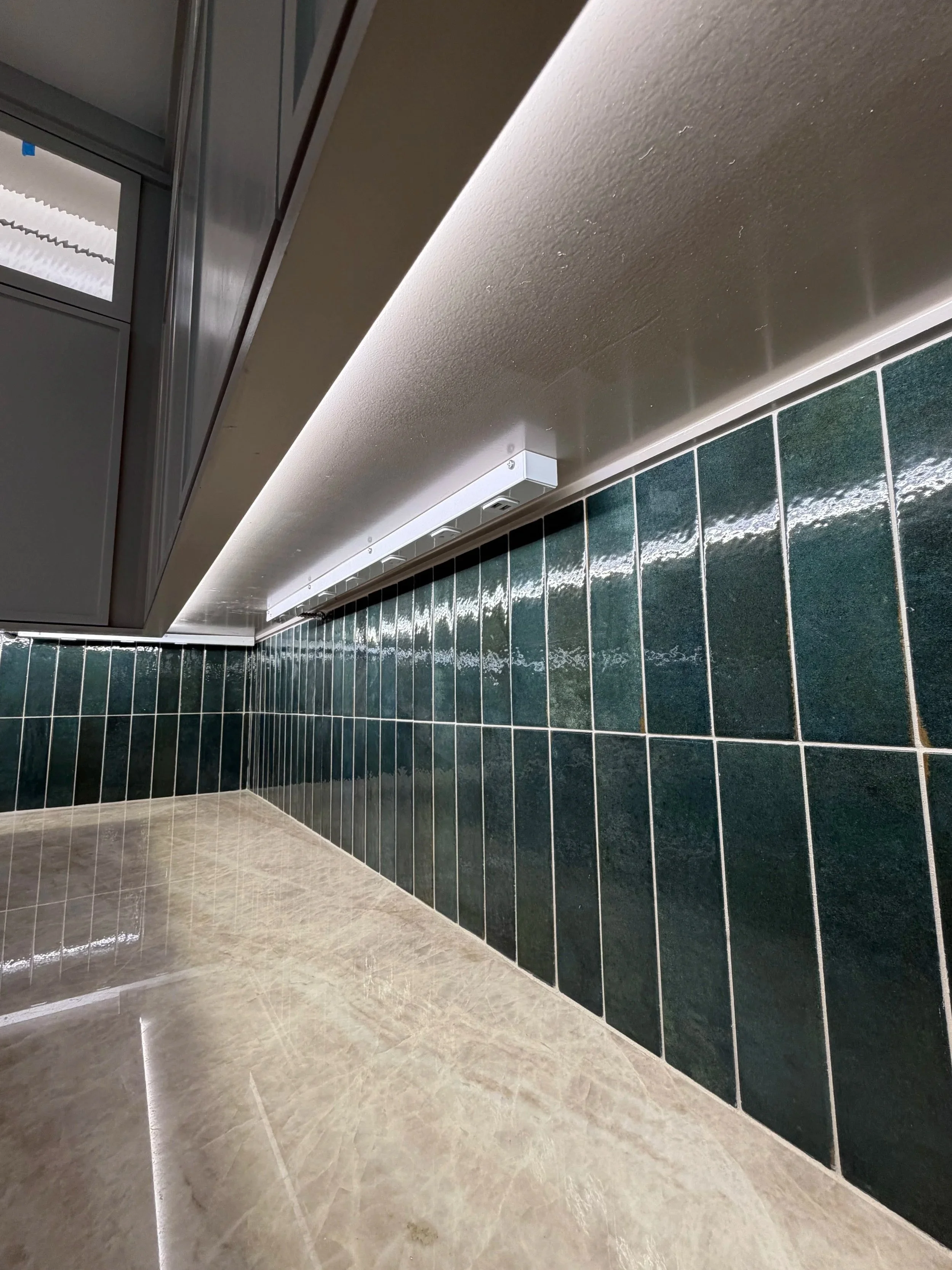 Close-up view of a kitchen countertop with dark green tiled backsplash and beige floor tiles, with a white power strip mounted under the upper cabinets.