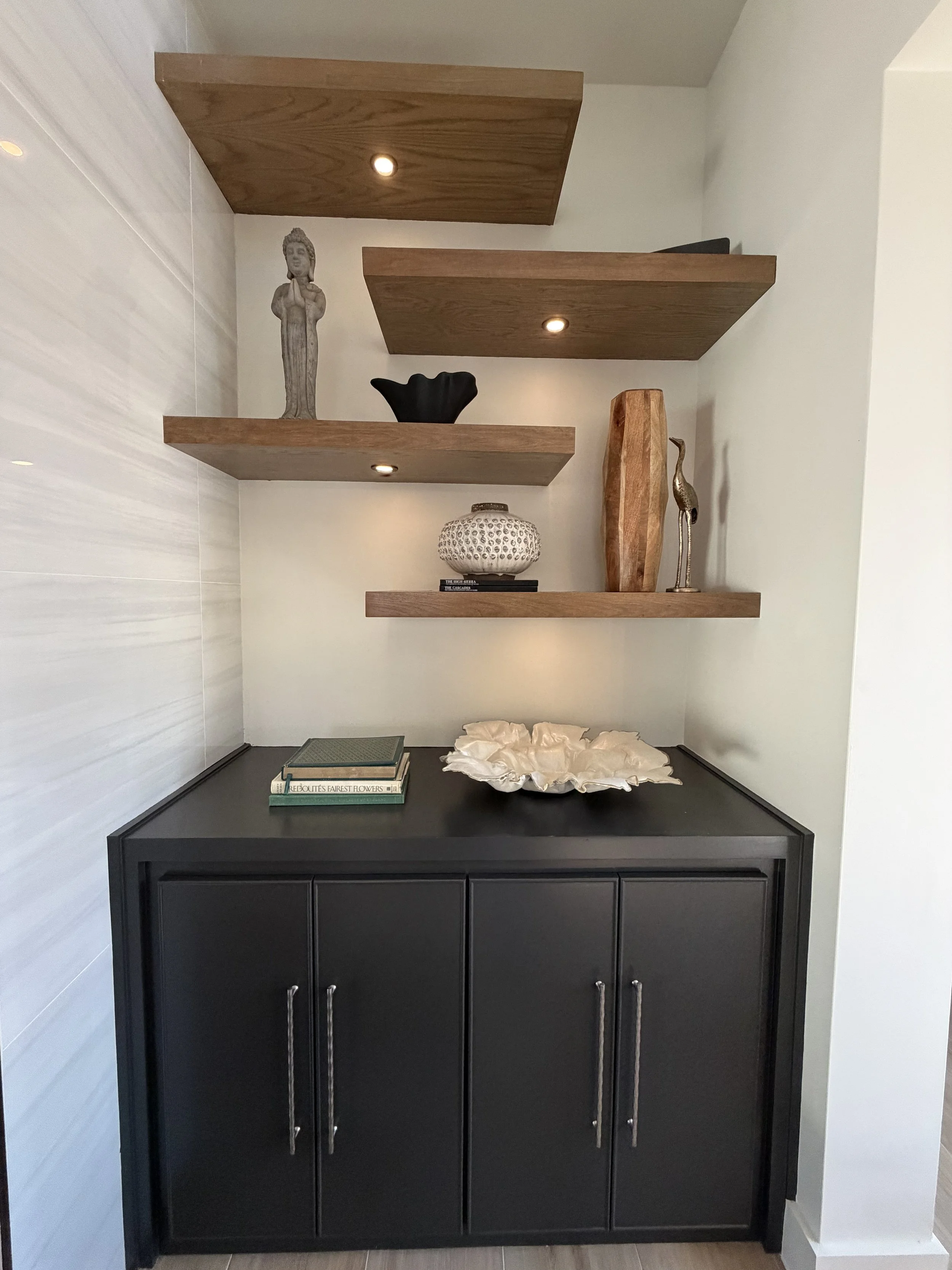 Black cabinet with four doors and silver handles topped with decorative objects, including books and a white fabric flower, against a light-colored wall with three wooden shelves holding various decorative items like statues, vases, and bowls.