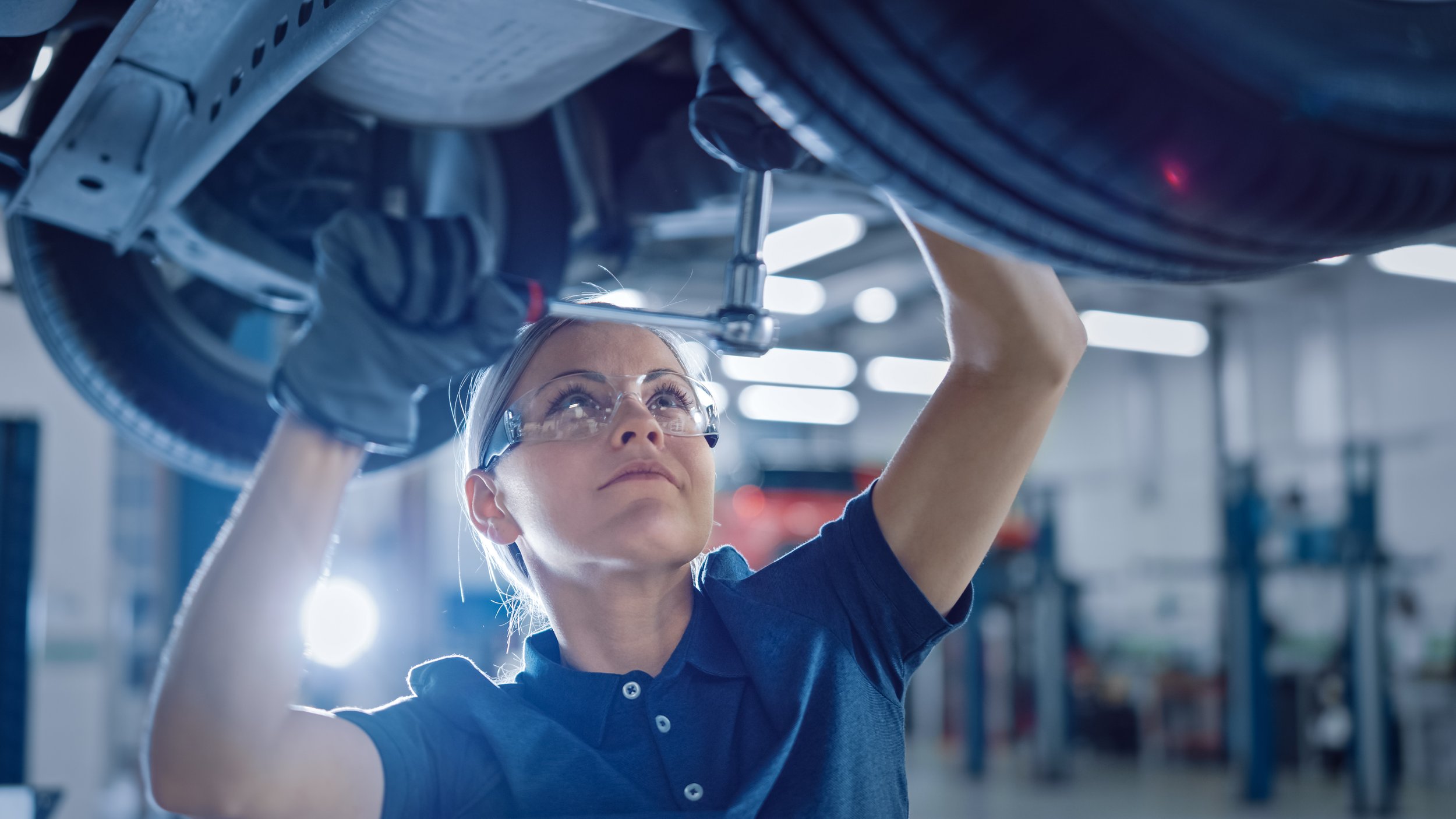 A female auto mechanic wearing safety glasses and gloves working under a car in a garage.