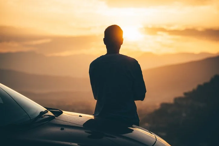 Person with crossed arms standing on a car, watching a sunset over mountains and clouds.