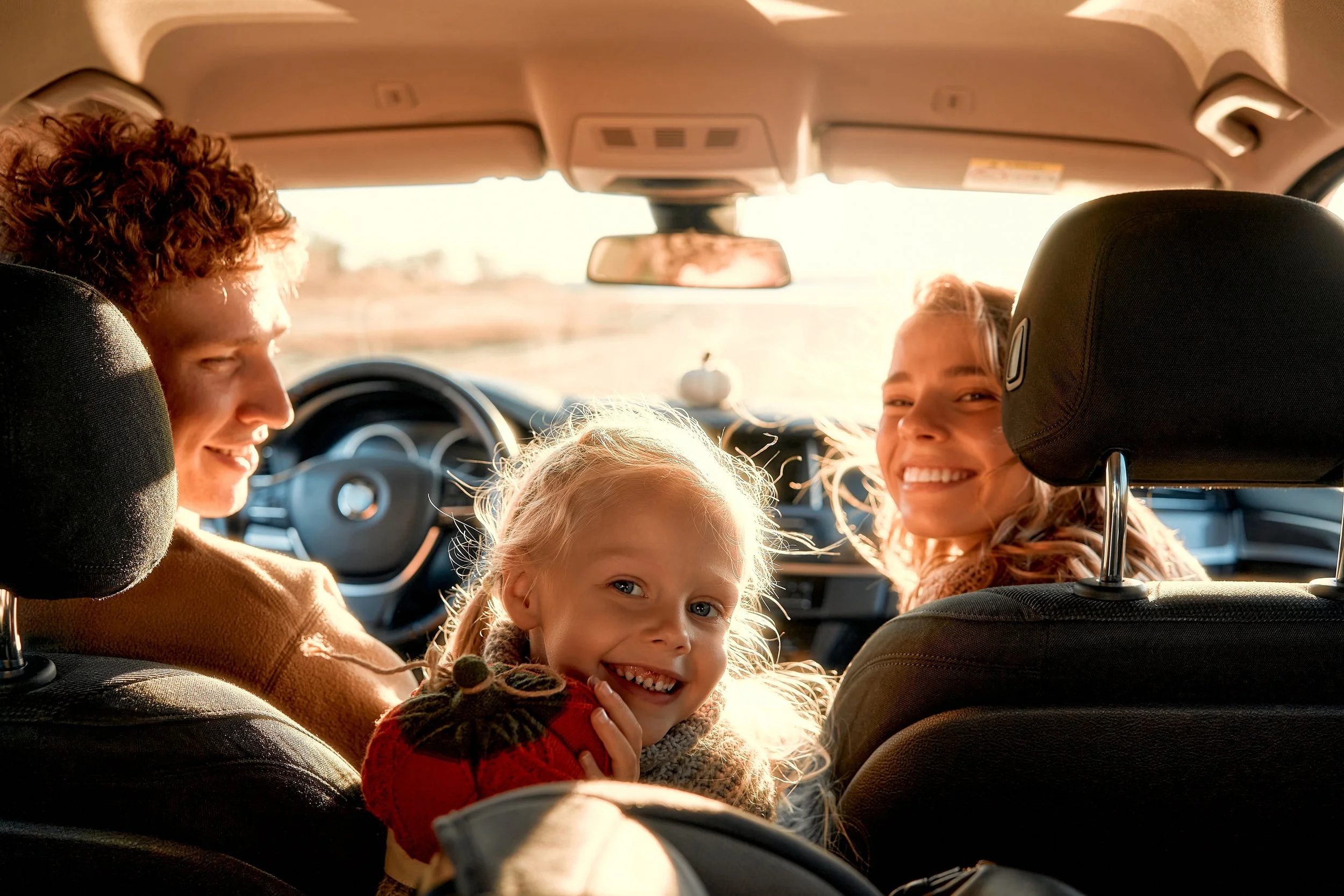 A family of four smiling inside a car during sunset, with the driver and passenger, a woman, visible, and two young girls in the backseat, one with blonde hair and a red sweater and the other with light hair and a gray scarf.