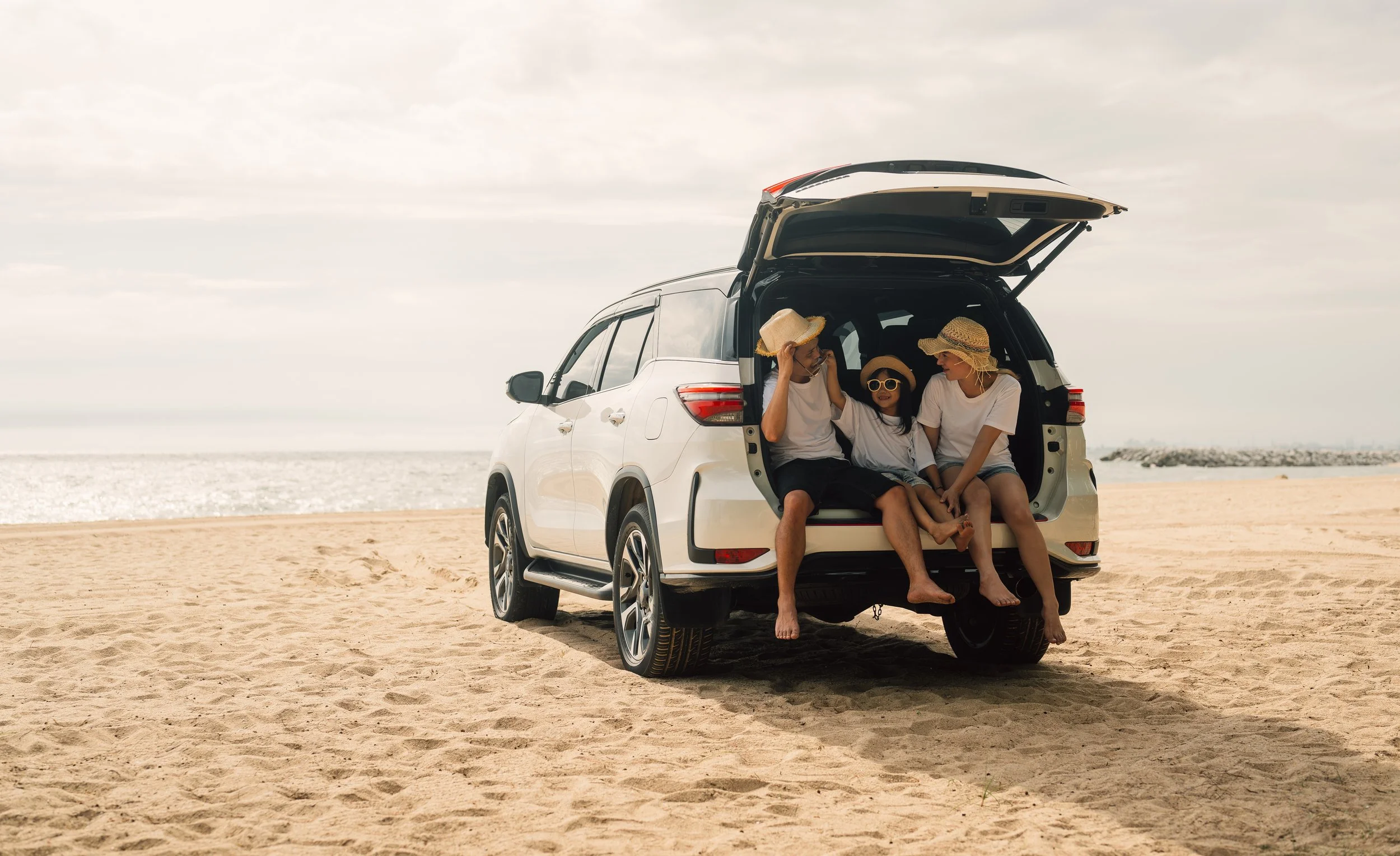 Family of three sitting in the open trunk of a white SUV on a sandy beach, wearing summer hats and sunglasses, with the ocean in the background.