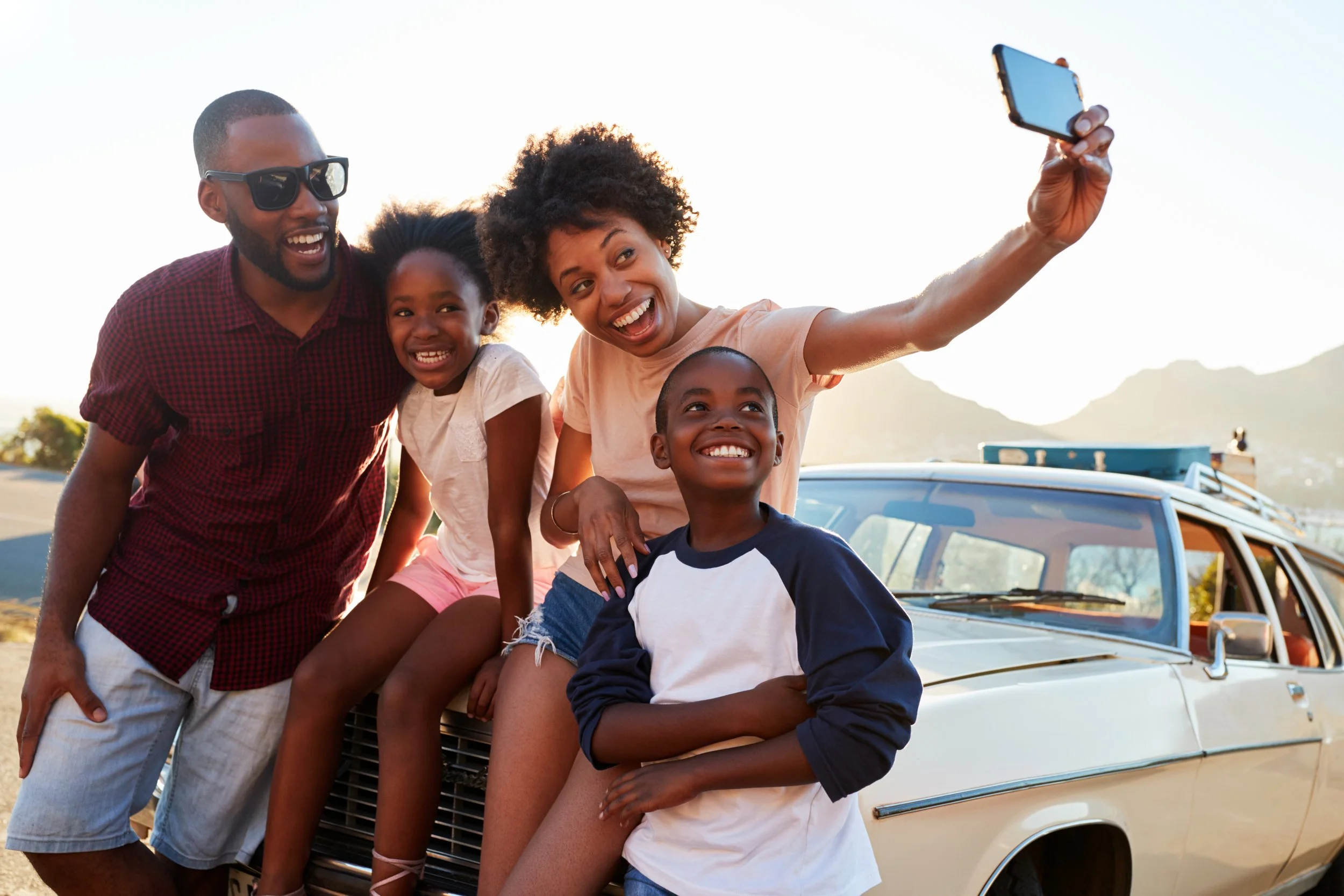 A smiling family taking a selfie outdoors near a vintage car during sunset, with mountains in the background.
