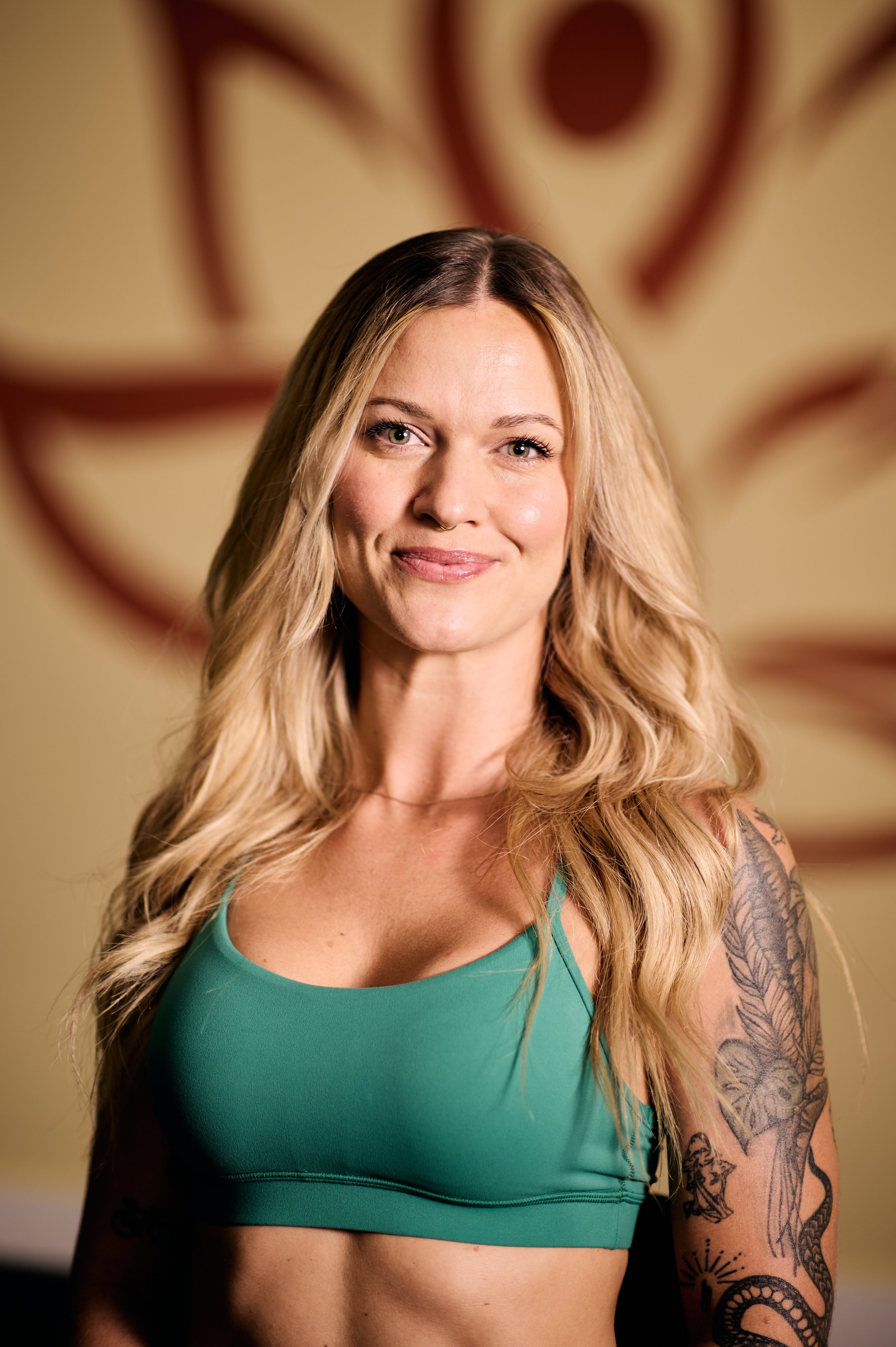 A woman with long blonde hair, wearing a teal sports bra, standing in front of a blurred background with a large, stylized eye design.