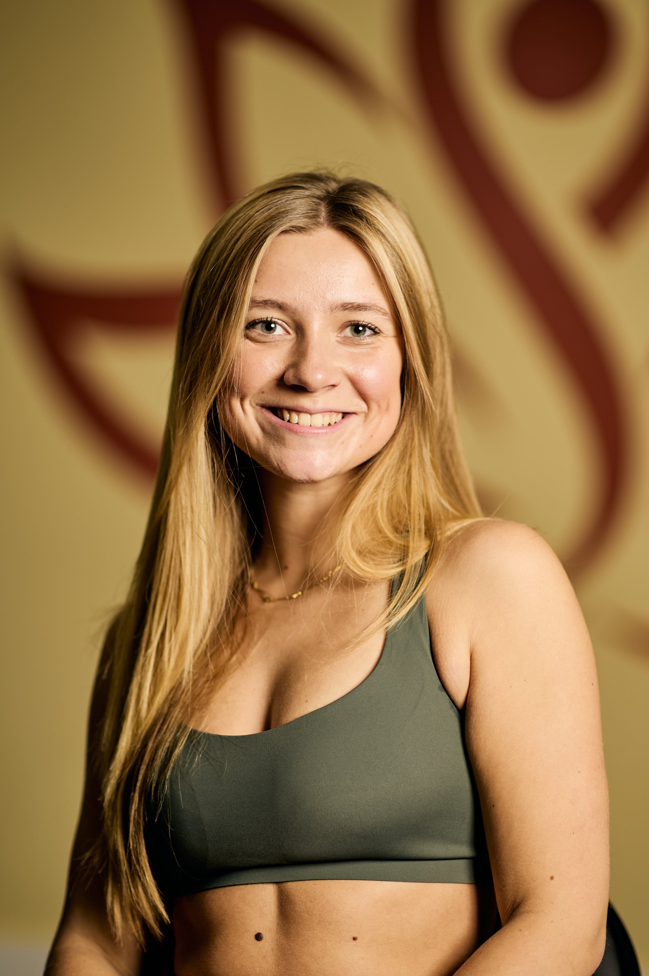A young woman with long blonde hair smiling in a gym or fitness setting, wearing a gray sports bra.