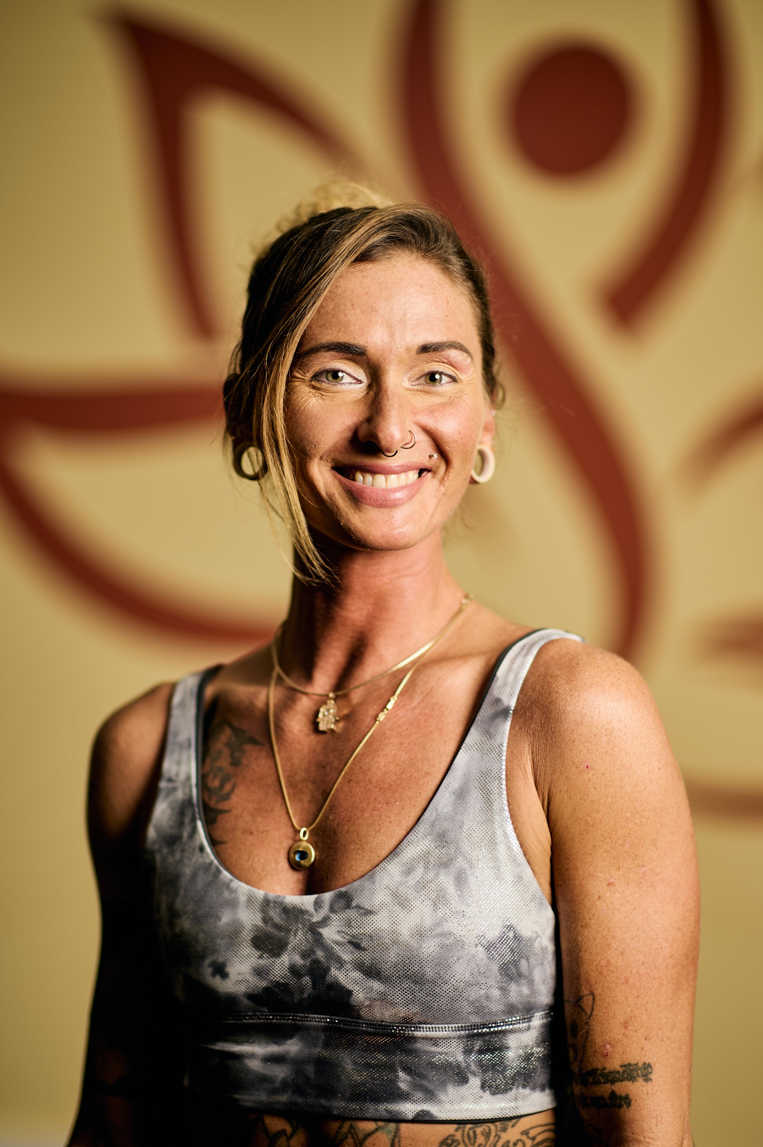 A smiling woman with a septum piercing, tattoos, tattoos, wearing a tie-dye crop top, and gold jewelry, standing in front of an abstract wall art.